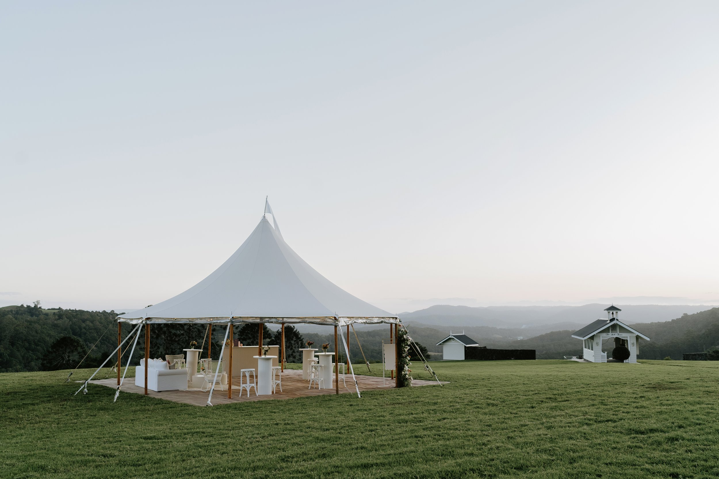 Large white event tent set up on a grassy field with outdoor furniture inside, surrounded by a scenic landscape of rolling hills and a small white building in the distance, under a clear sky.