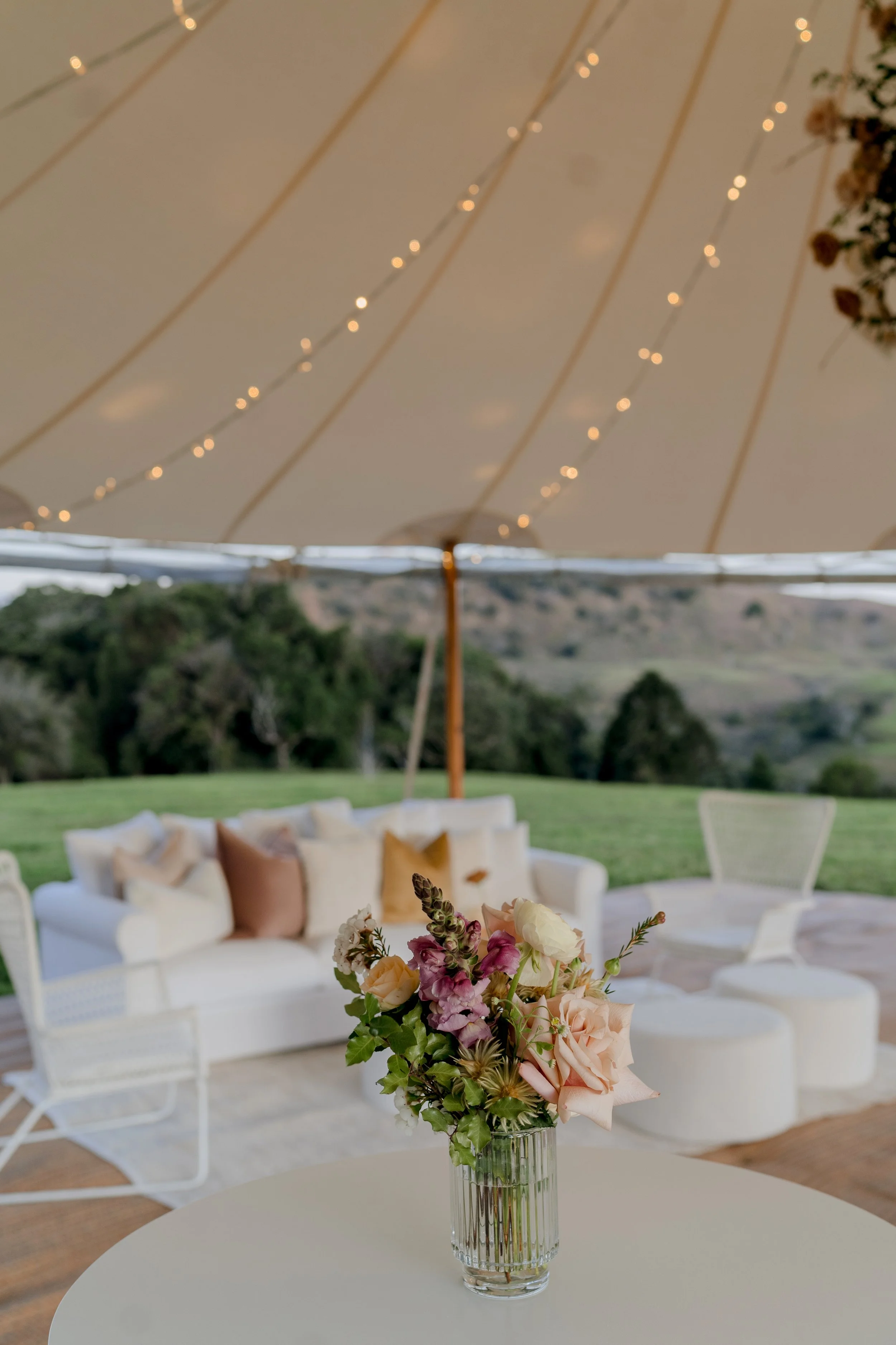Elegant outdoor event setup with a floral bouquet in a glass vase on a round table, white sofa with beige and brown cushions, white chairs, and a large tent with string lights, overlooking a green landscape.