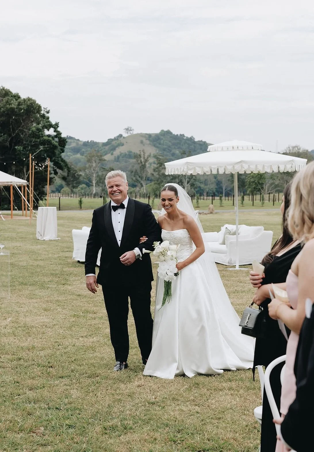 A bride walking happily arm-in-arm with an older man, likely her father, during a wedding ceremony outdoors on a grassy field with hills in the background. The bride wears a white strapless wedding gown and holds a bouquet of white flowers. The man is in a black tuxedo with a bow tie, smiling widely. There are white furniture and parasols on the field, with a few guests visible on the side.