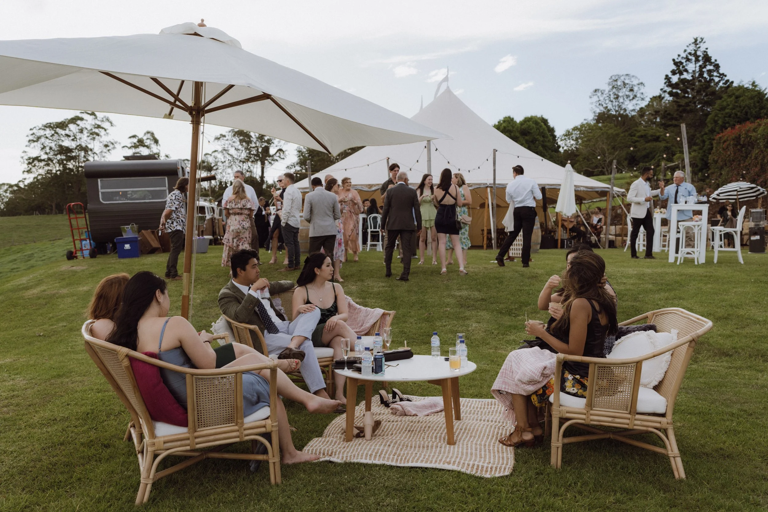 People socializing and relaxing outdoors at a garden party or wedding with a large white tent, umbrellas, and outdoor furniture on a grassy area.