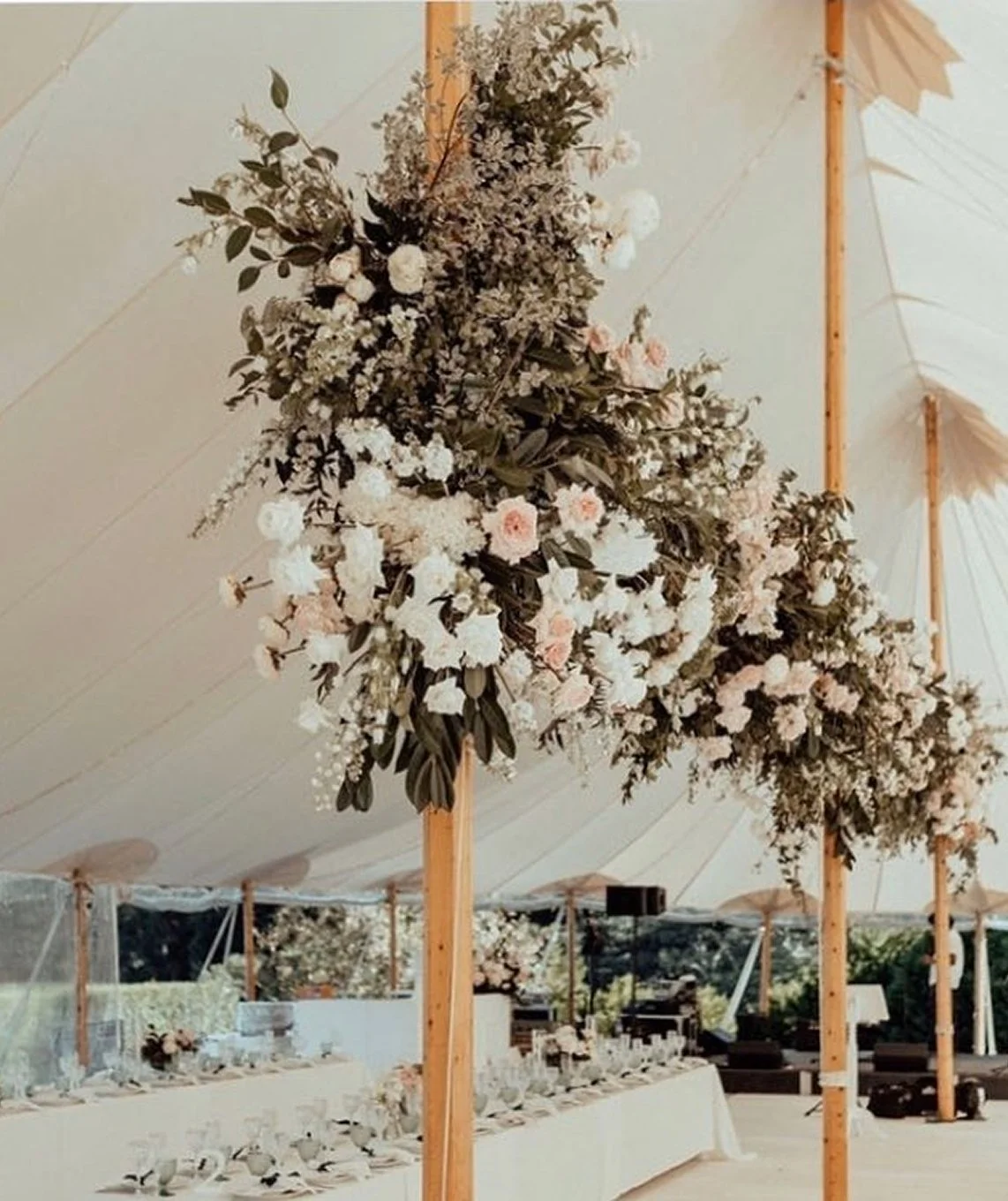Elegant outdoor wedding reception decorated with tall floral arrangements of white and blush roses, greenery, under a large white tent with wooden support poles.