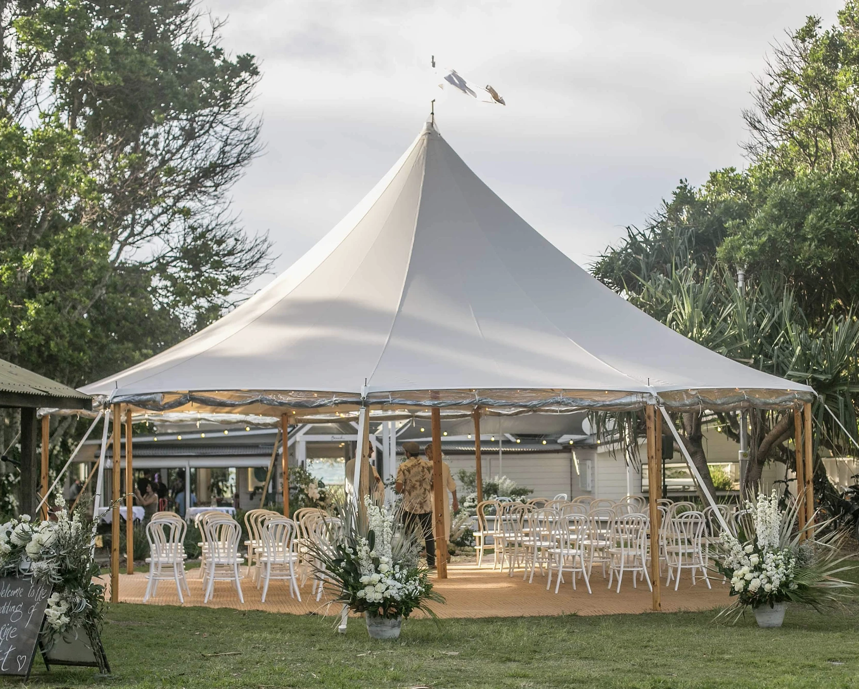 A large outdoor white wedding tent decorated with floral arrangements, with white chairs arranged inside for the ceremony.