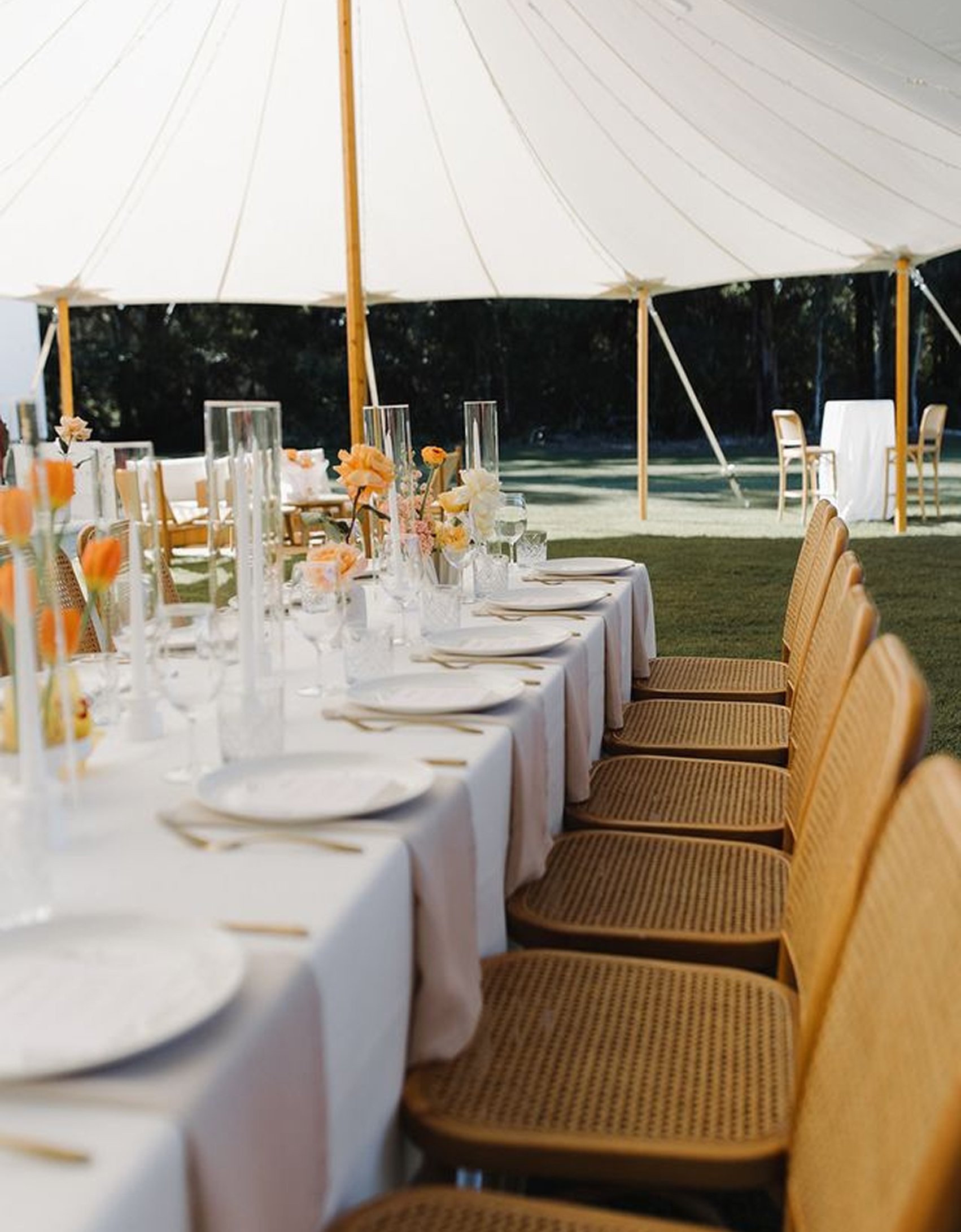 Elegant outdoor dining setup with a long table covered in a white tablecloth, decorated with pink and orange flowers in vases, tall glass candle holders, plates, glasses, and cutlery, set under a large white canopy, with wooden chairs arranged around the table and additional tables and chairs in the background.