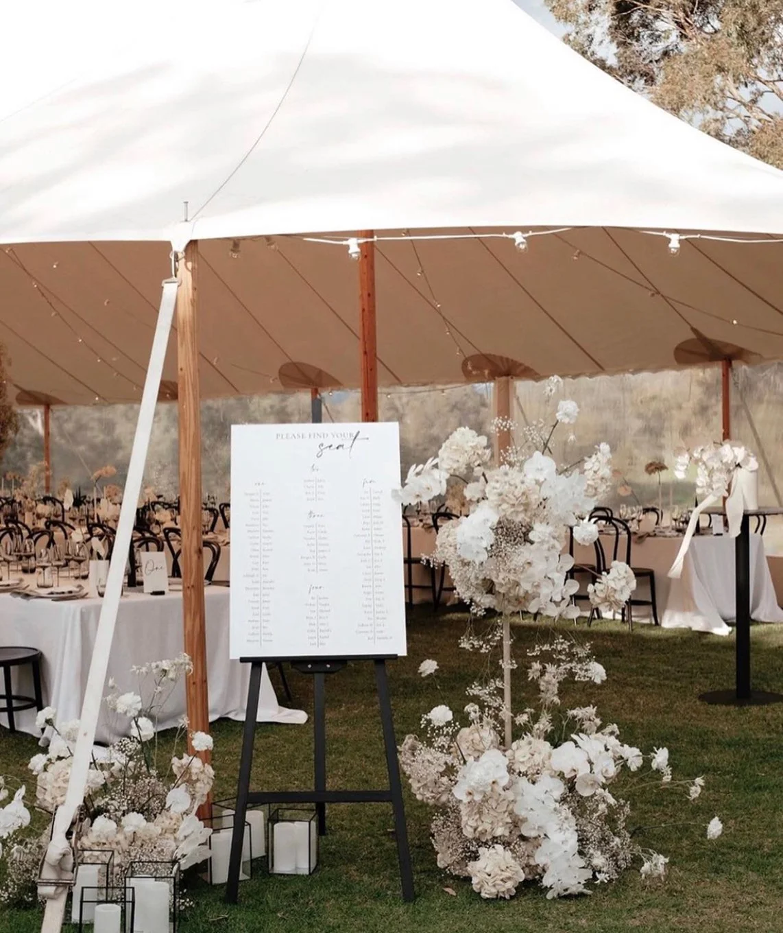 Wedding reception setup with long tables, floral arrangements, and a seating chart stand under a large canopy.