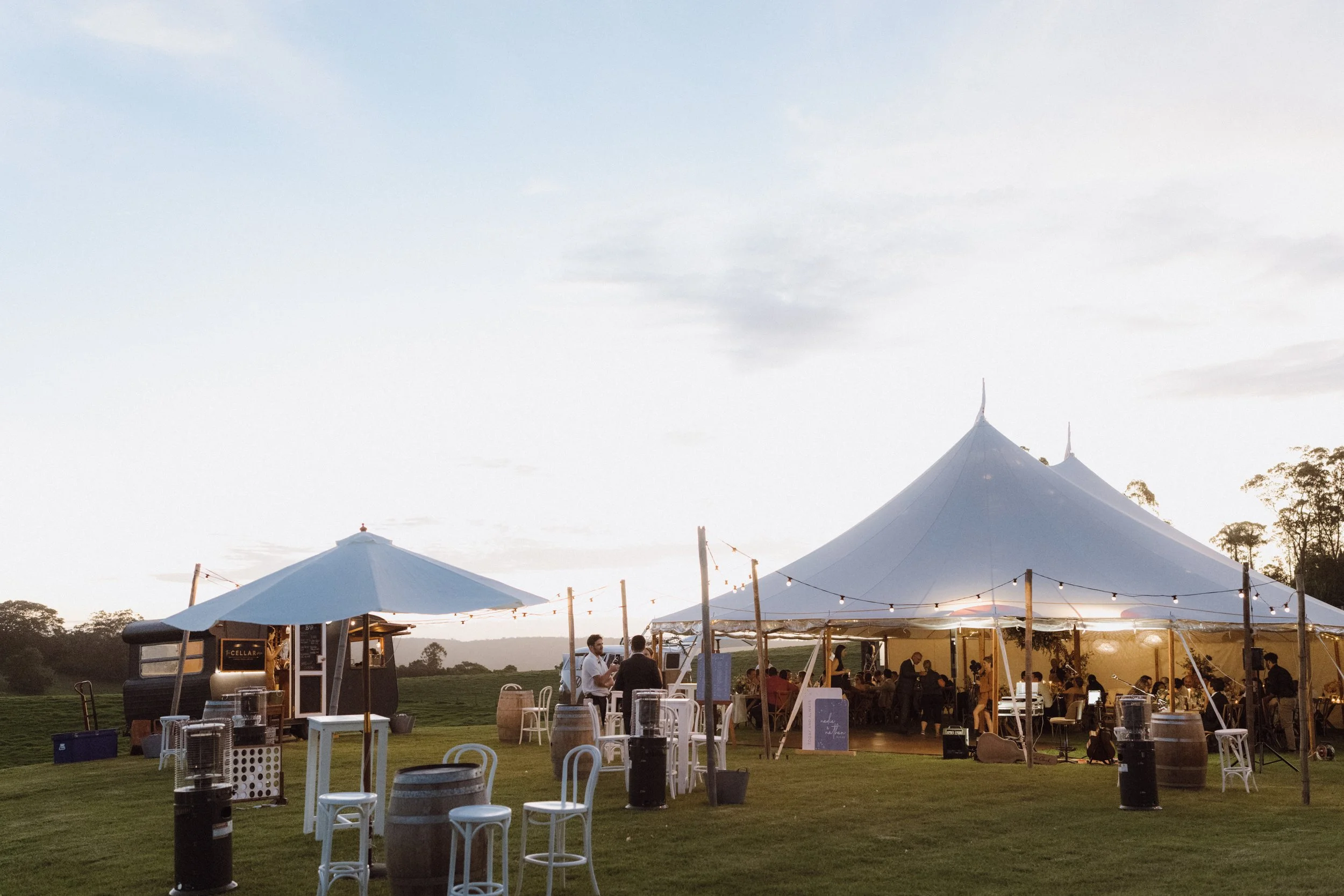 Outdoor event with large white tent, string lights, smaller tent, and people sitting and standing around on grass field during sunset.