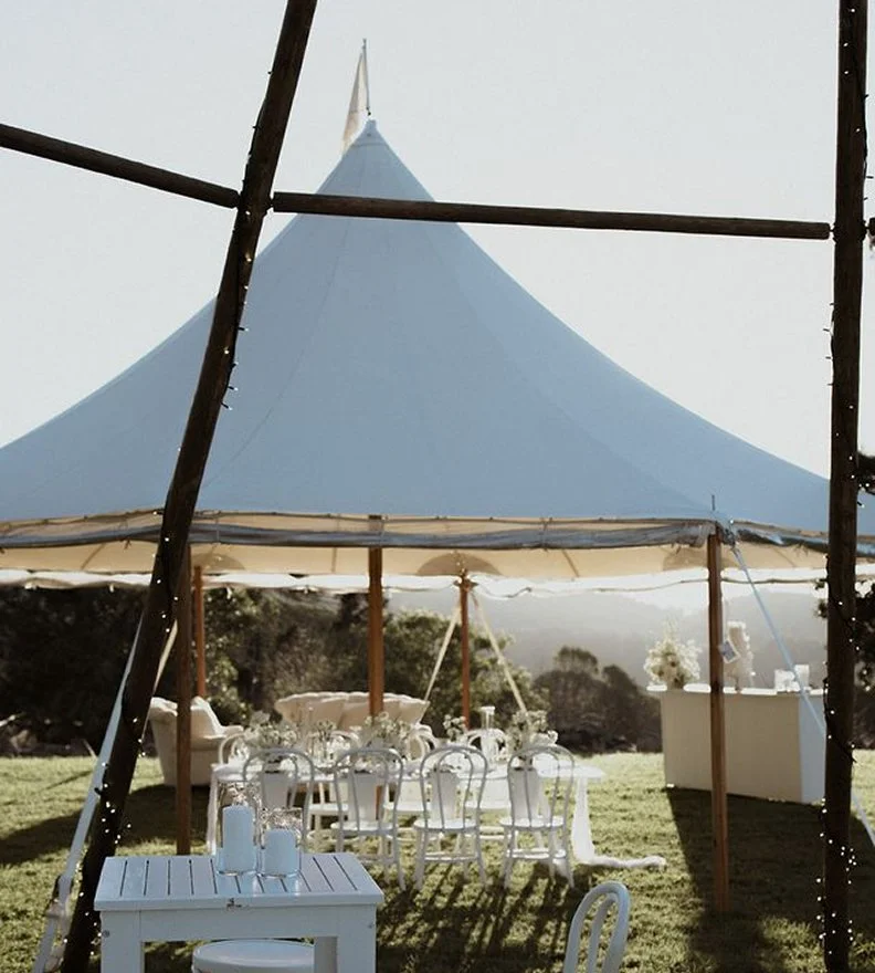 Outdoor event setup under a large white tent with tables, chairs, and floral decorations on a grassy field.