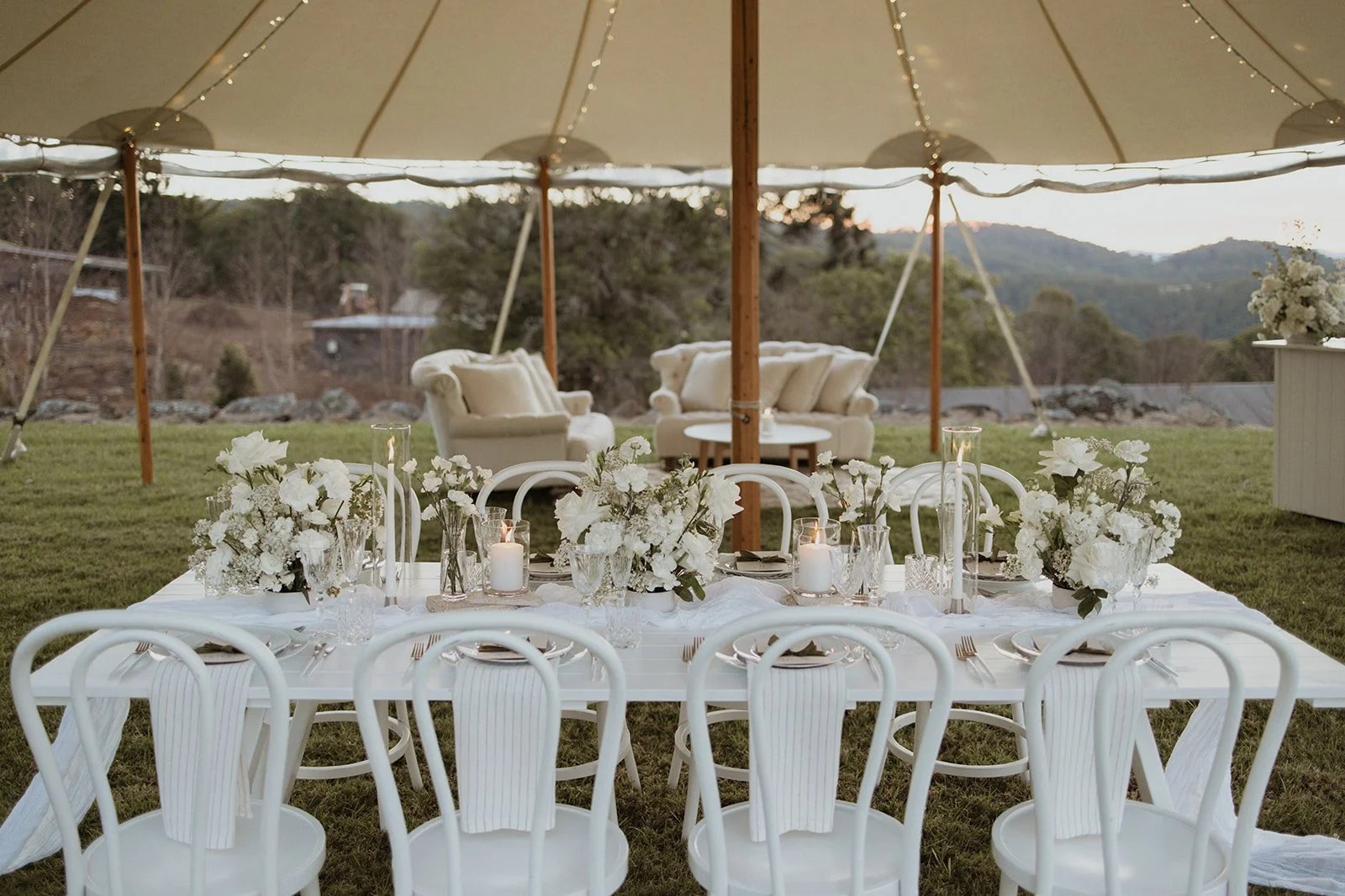 Elegant outdoor event setup with a white rectangular table decorated with white floral arrangements, candles, and glassware, under a large tent with a scenic mountain view in the background.