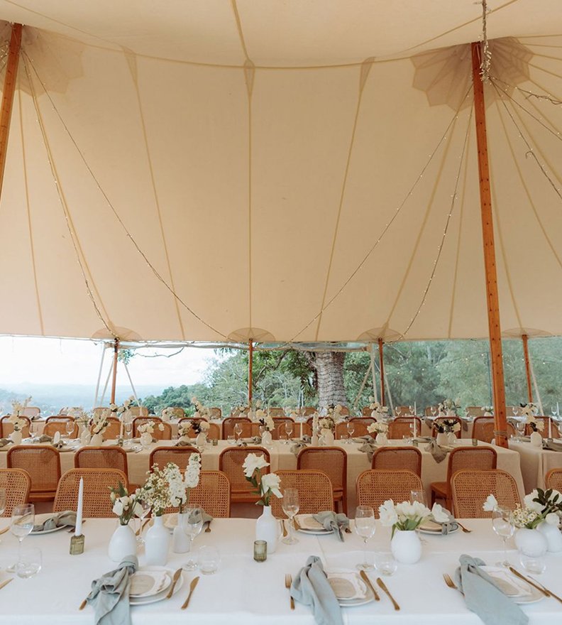 Long banquet tables set with white vases filled with white flowers, glasses, plates, napkins, and cutlery under a large beige tent with wooden supports, in an outdoor setting with trees visible in the background.