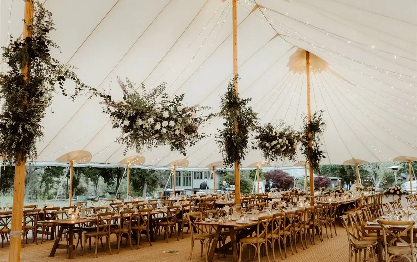 Elegant outdoor wedding reception under a large white tent with hanging floral arrangements and string lights, wooden tables and chairs set for guests.