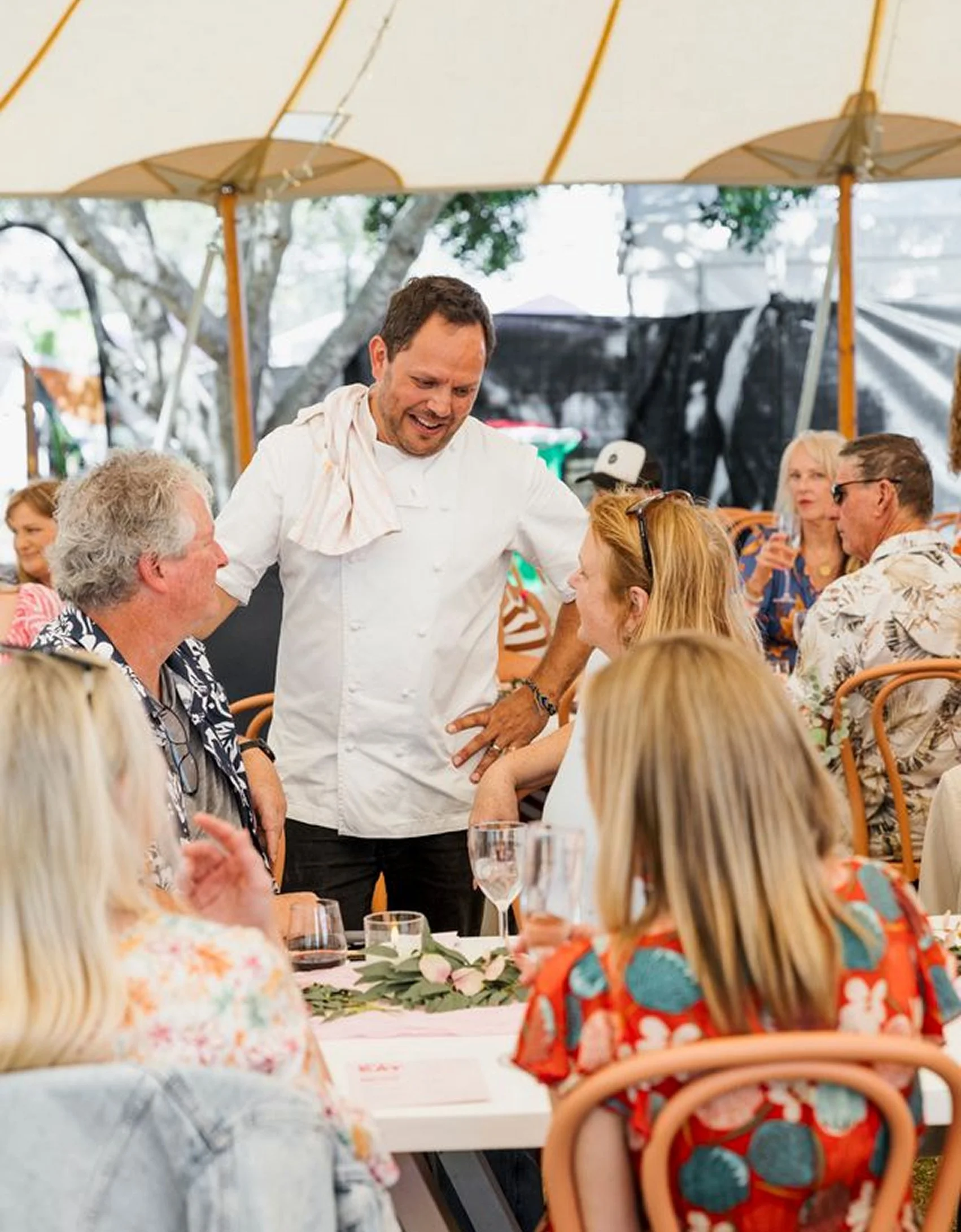 Chef talking and smiling with a woman at a table during a gathering or celebration facing a large tent outdoor.