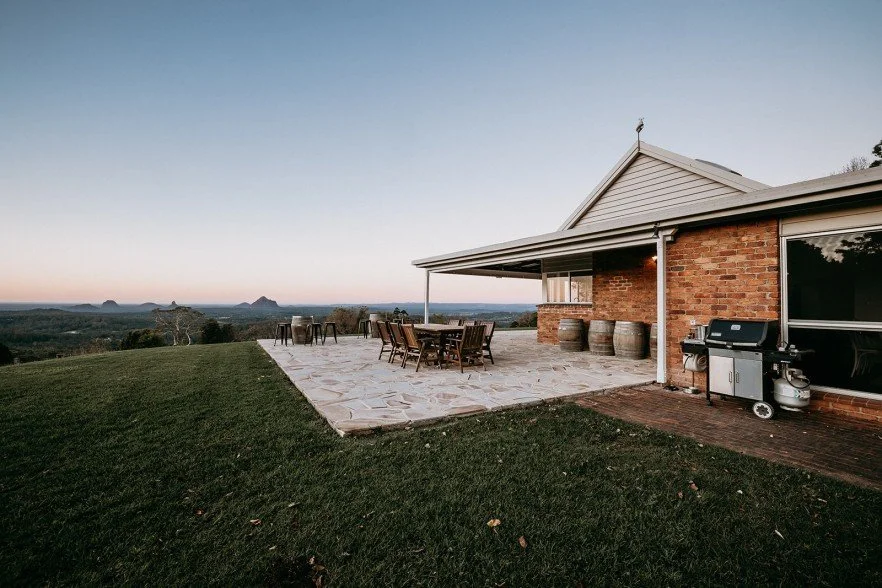 Backyard patio with outdoor dining table, chairs, and a barbecue grill, overlooking a scenic landscape with hills and a clear sky.