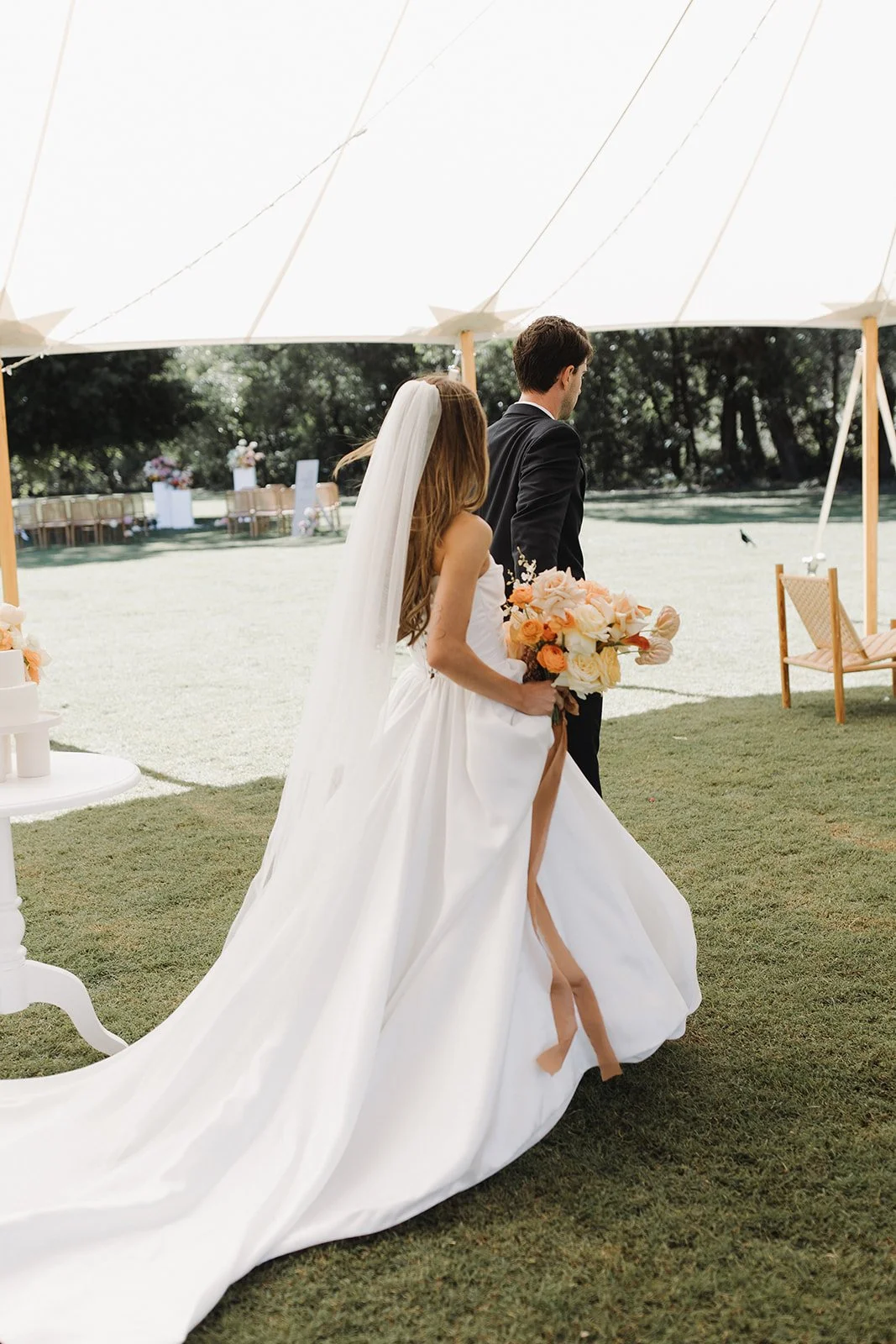 Bride and groom walking under a tent at an outdoor wedding, the bride holding a bouquet of peach and white flowers, wearing a white gown with a long train and veil, the groom in a black suit.