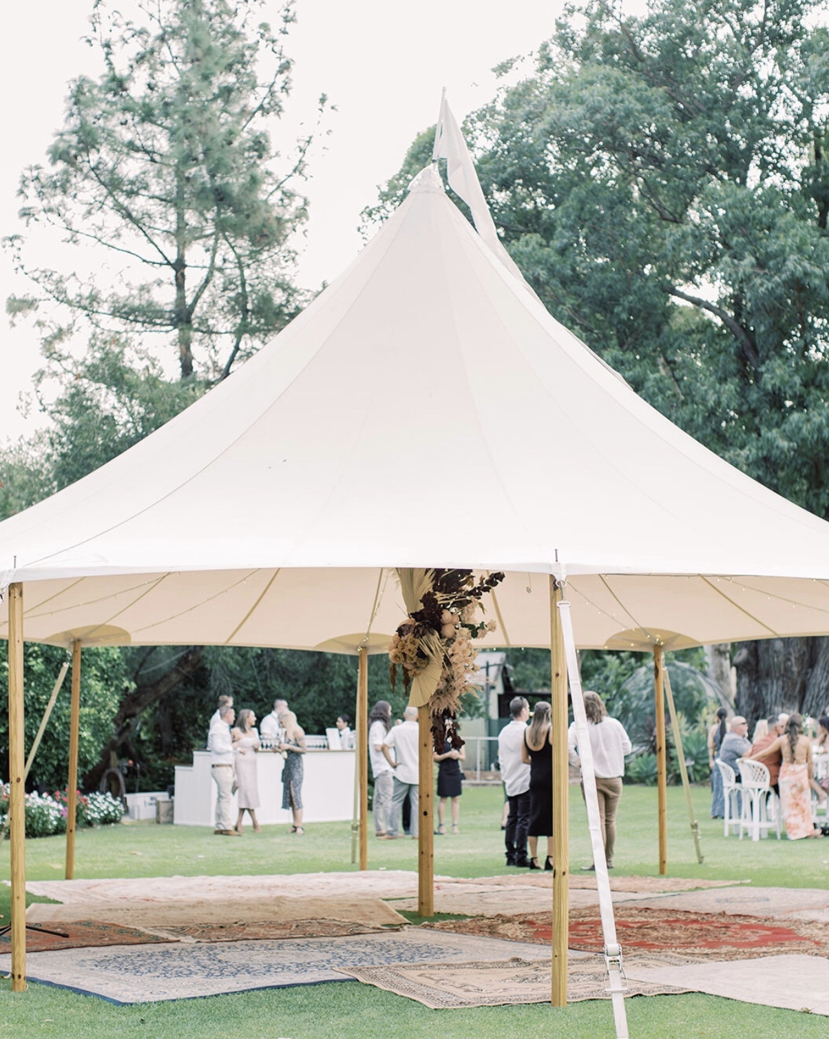 Outdoor event under a large white tent with people socializing on a grassy lawn surrounded by trees, decorated with a flower arrangement hanging from the tent's support pole.