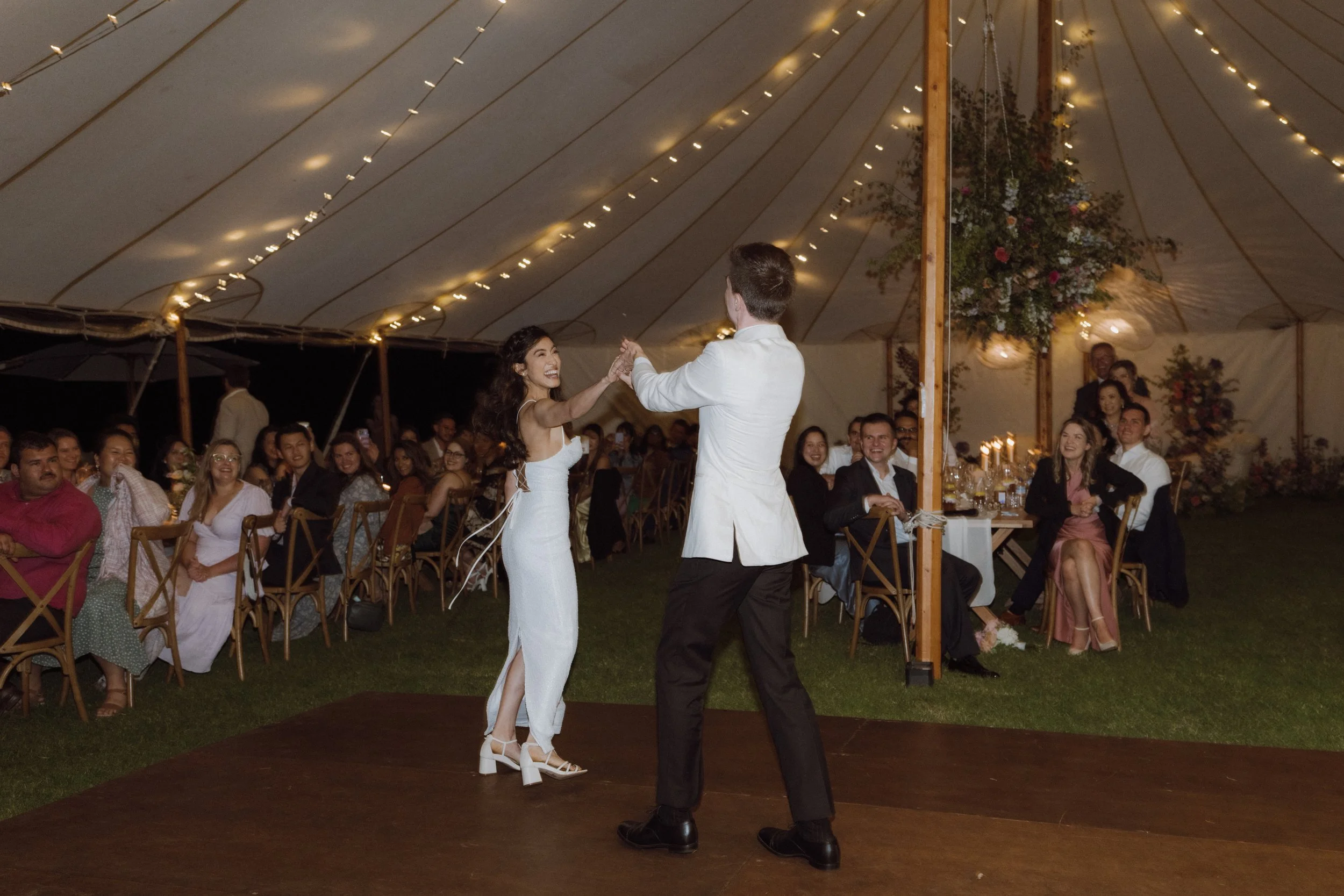 A couple dancing at a wedding reception under a large decorated tent with guests watching and smiling.