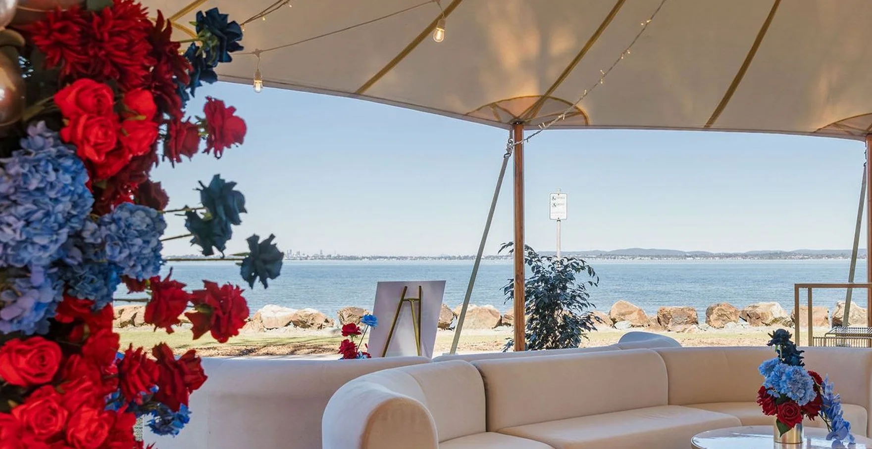 Outdoor seating area with a white sofa decorated with flowers, overlooking a body of water with rocks and a distant shoreline under a canopy with string lights.