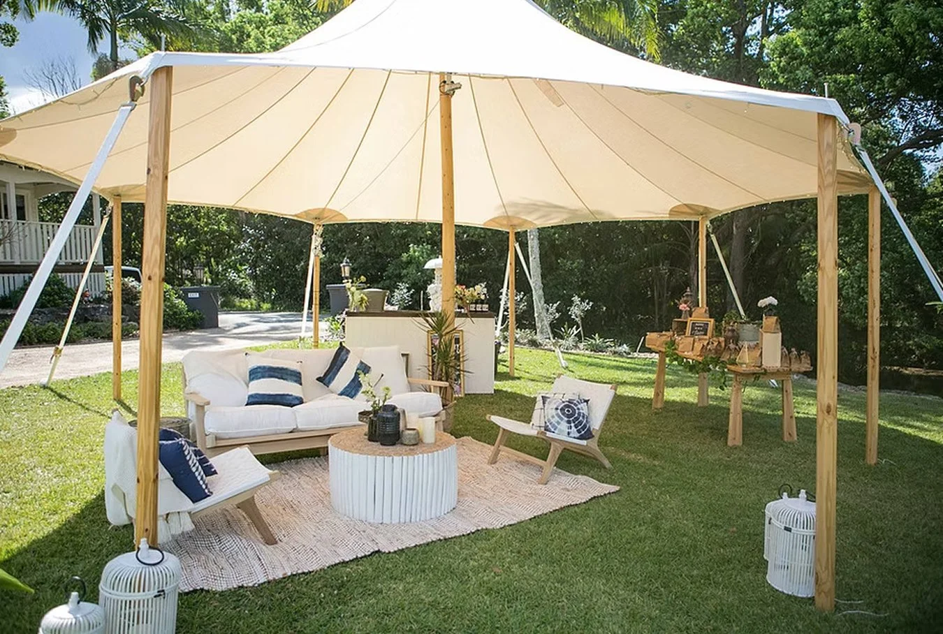 Outdoor seating area under a large beige canopy with white and navy blue cushions, small tables, and decorative items on a grassy lawn surrounded by trees.