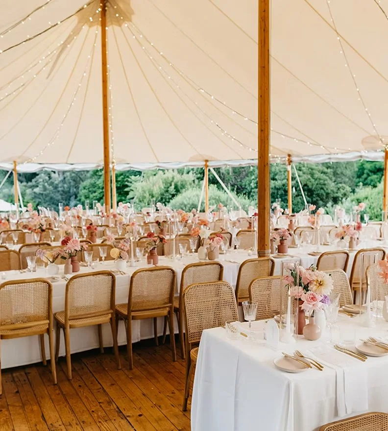 Decorated outdoor event tent with wooden poles, string lights, and a long table set with pink and white floral arrangements, glassware, and gold cutlery, overlooking a green landscape.