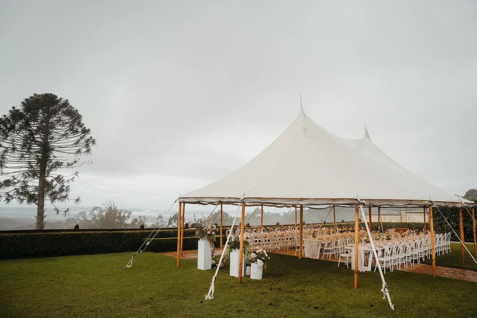 White event tent with wooden poles, set up outdoors on a lush green lawn, decorated with flowers, under a cloudy sky.
