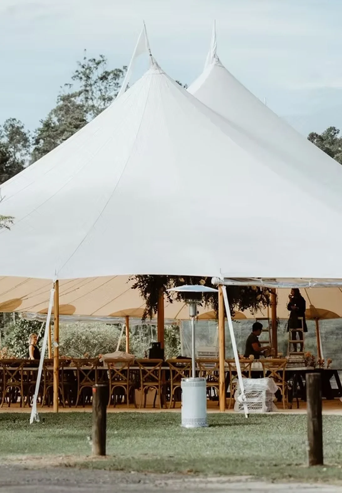A large white tent with a high peaked roof set up outdoors, with tables and chairs underneath it, and a person working on a ladder inside.