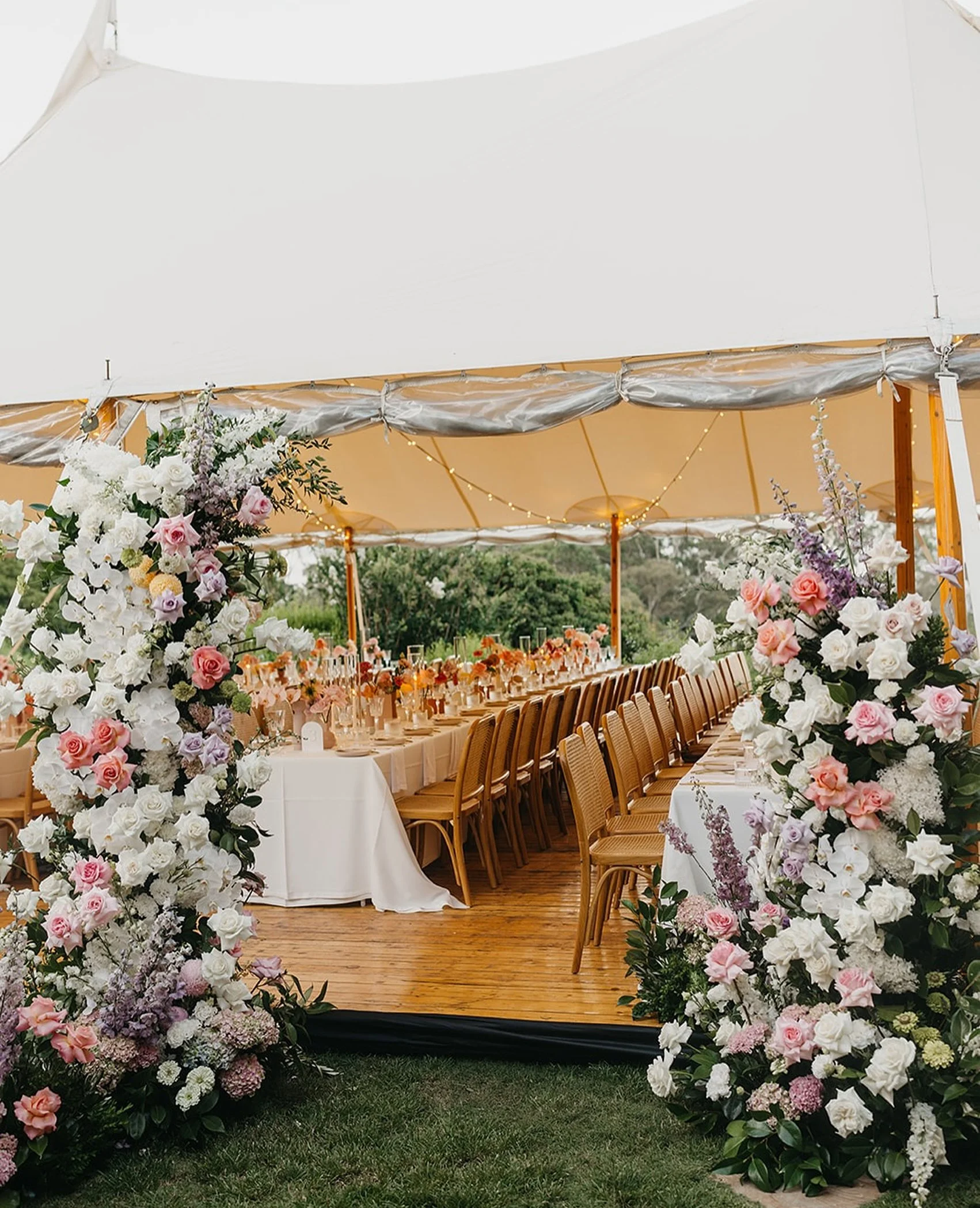 Elegant outdoor wedding reception setup under a large white tent with a long decorated table, surrounded by floral arrangements of pink, white, and purple flowers, and wooden chairs.