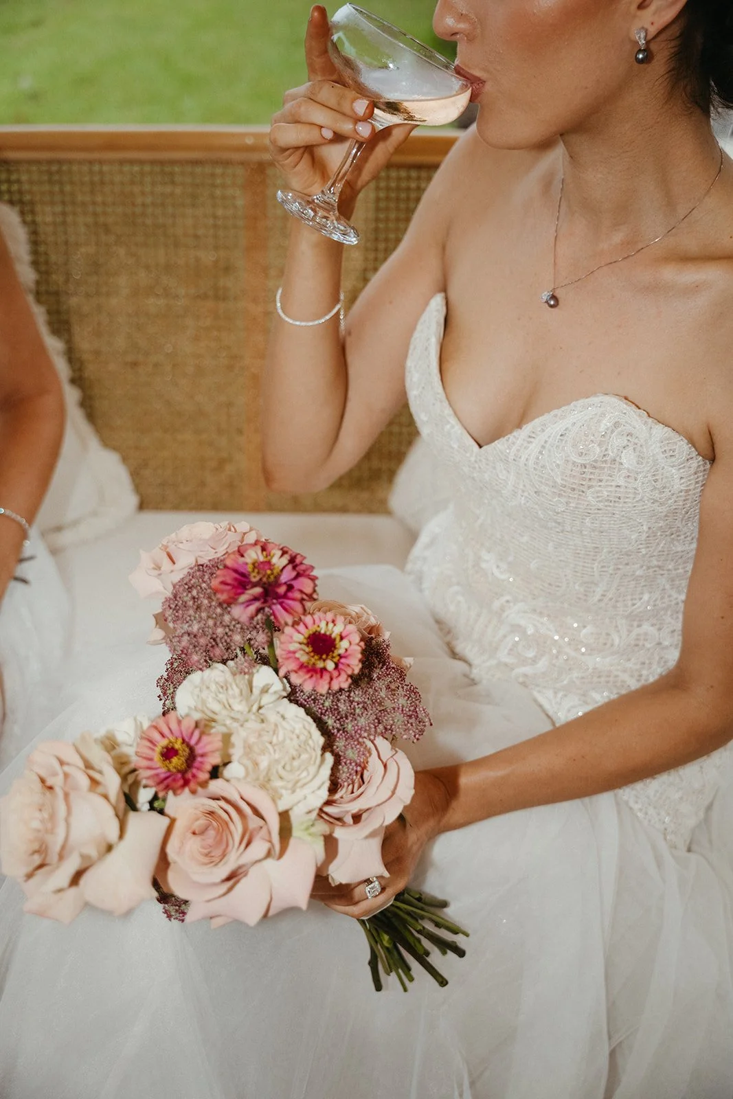 Woman in a wedding dress holding a bouquet of pink and white flowers, sipping from a glass of wine.