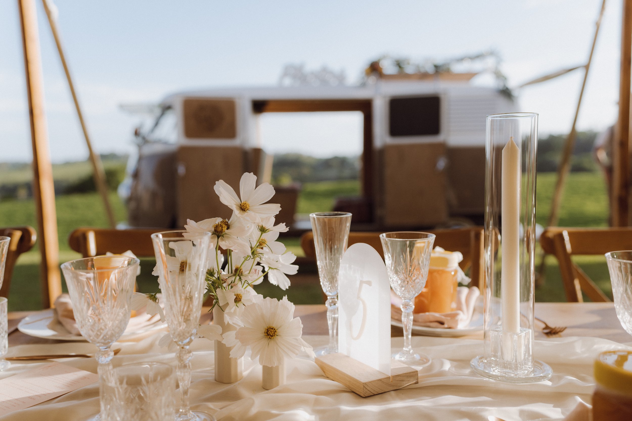 Table decorated with white flowers, glassware, and a candle, set outdoors with a camper van in the background.