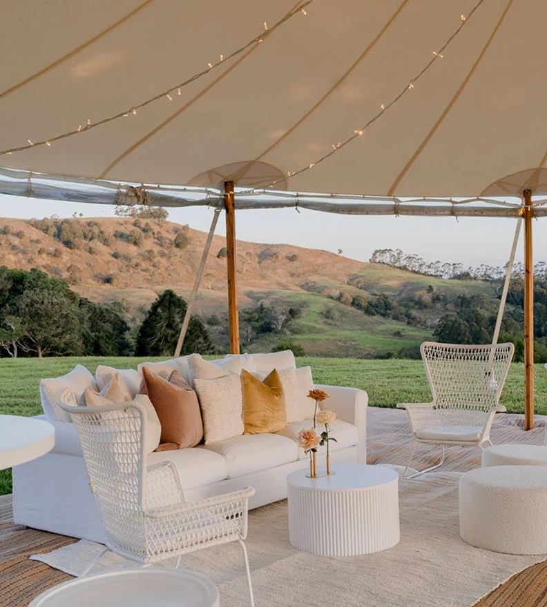 Outdoor seating area under a large beige tent with white wicker chairs, a white couch with pillows, and a small round table with flowers. Green rolling hills and trees in the background.