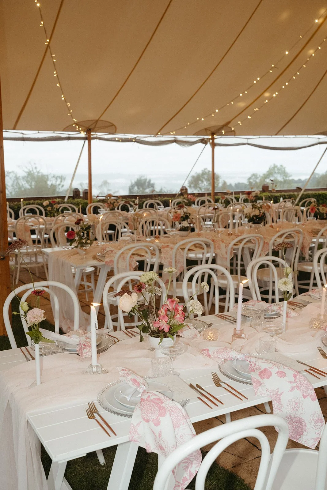 Decorated outdoor wedding reception with white tables, pink floral centerpieces, white chairs, candles, and string lights under a large tent.