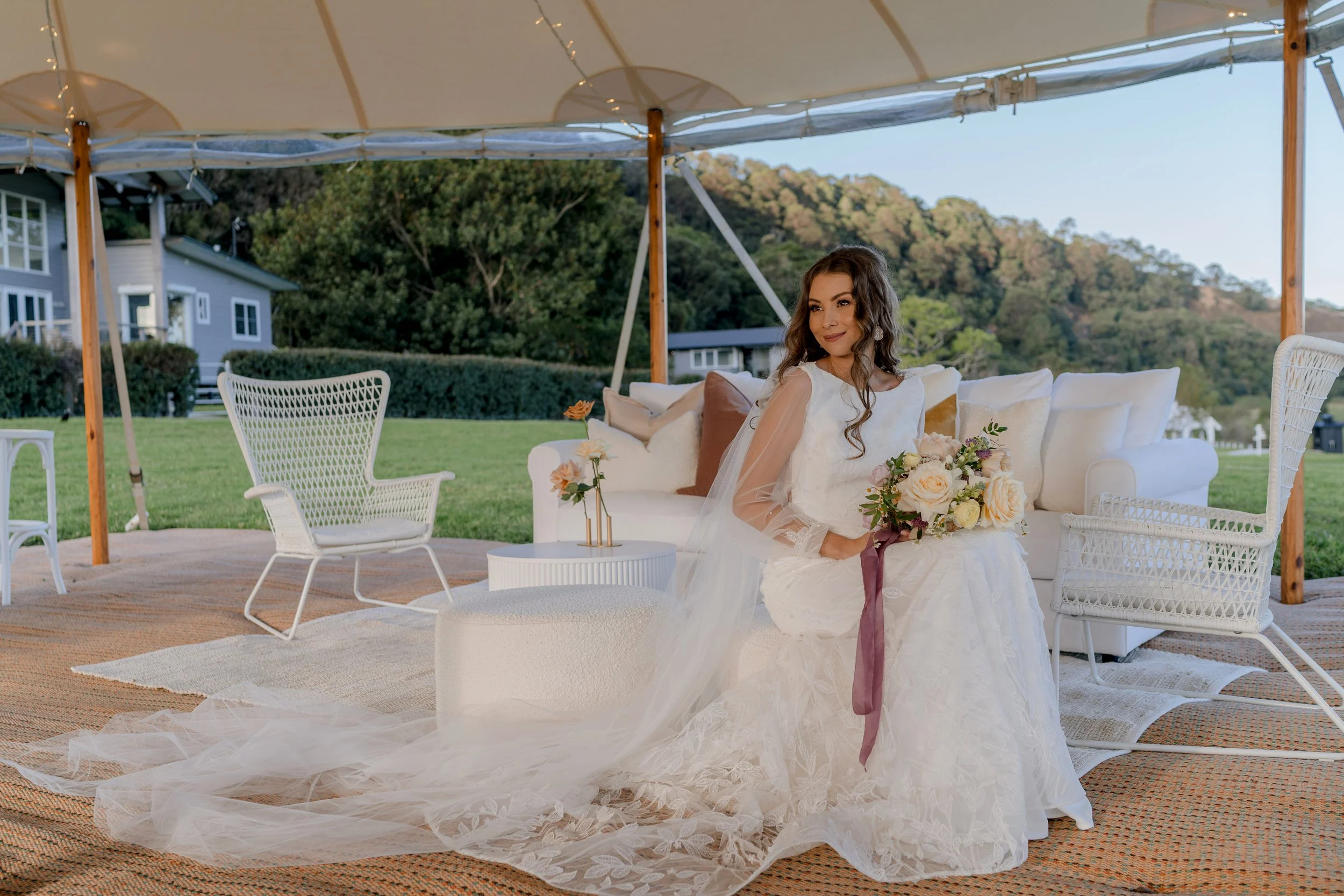 Bride in white wedding dress sitting on white ottoman holding a bouquet of flowers outdoors under a large tent. There are white chairs, a sofa, and a small table with flowers around her, with a green lawn and hills in the background.