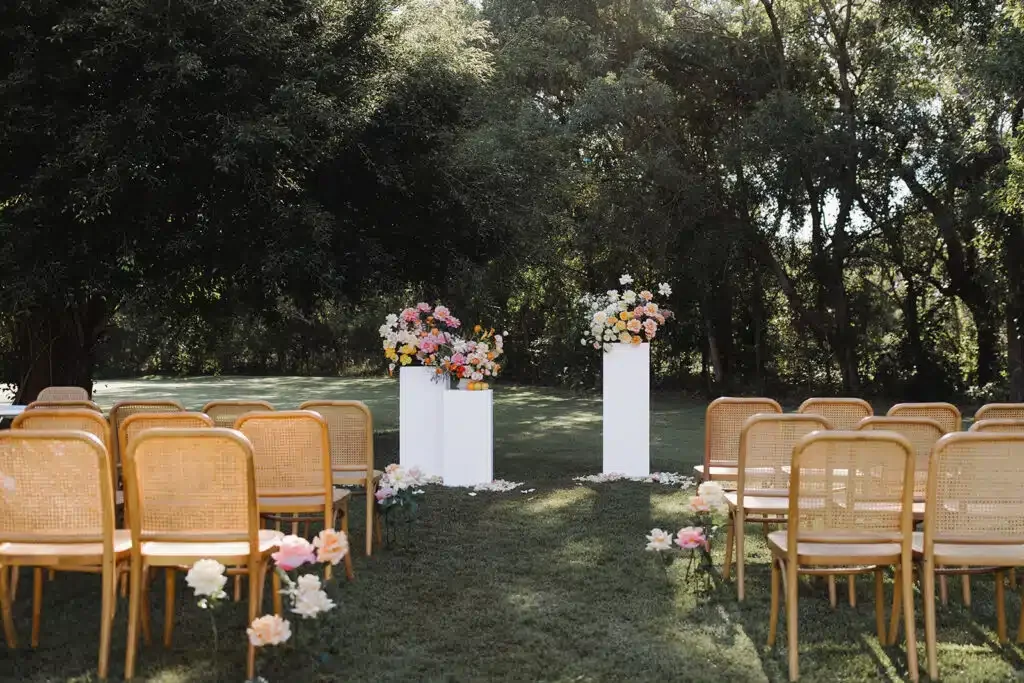 Outdoor wedding ceremony setup with chairs arranged on each side facing two white pedestals decorated with pink and white flowers, surrounded by trees and greenery.