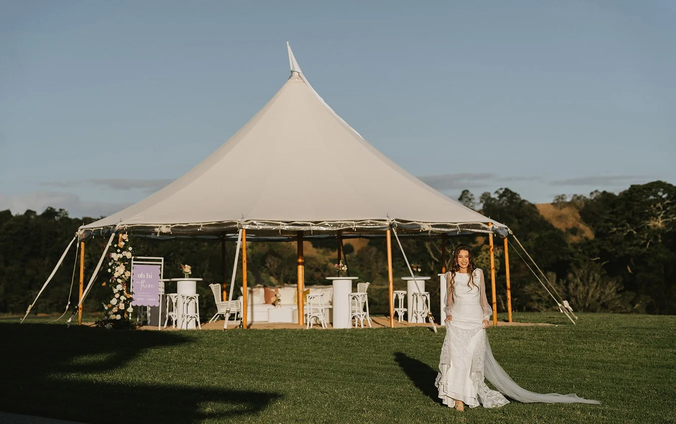 A woman in a white wedding dress standing on grass in front of a large white wedding tent decorated with flowers, with outdoor furniture inside, on a sunny day with a partly cloudy sky and trees in the background.