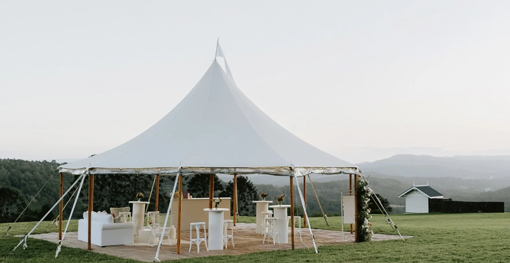 A white wedding tent set up outdoors on a grassy field with mountains in the background, decorated with white furniture and floral arrangements.