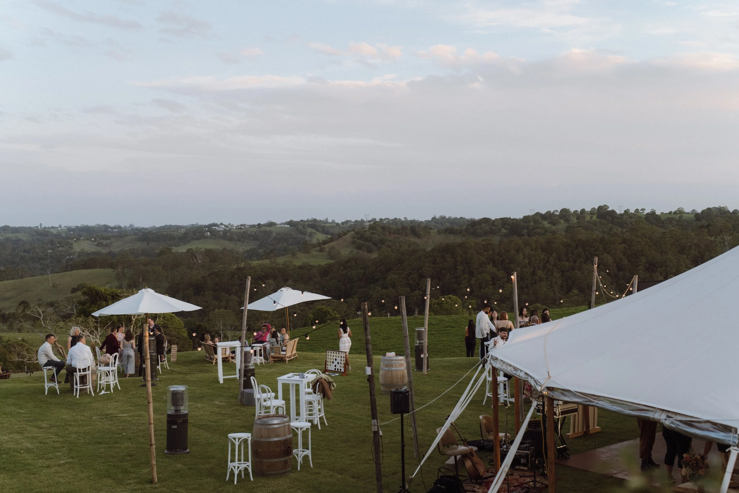 Outdoor wedding reception setup with white tents, tables, and chairs on a grassy hillside under a cloudy sky, with guests mingling and scenic rolling hills in the background.