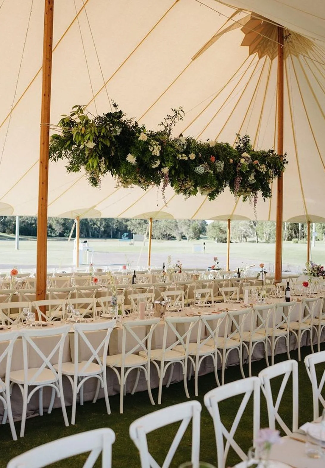 Long banquet table with white chairs and floral centerpieces under a white tent with hanging greenery and flowers, outdoor setting with trees in the background.