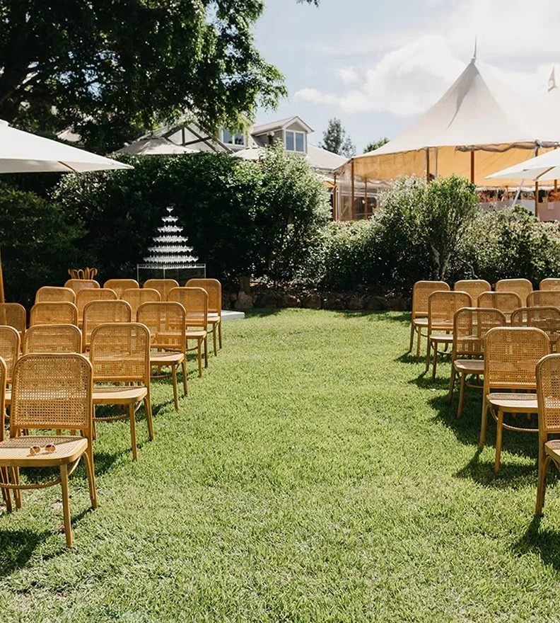 Outdoor wedding or event setup with rows of wooden chairs facing a water fountain, with a large tent and greenery in the background.