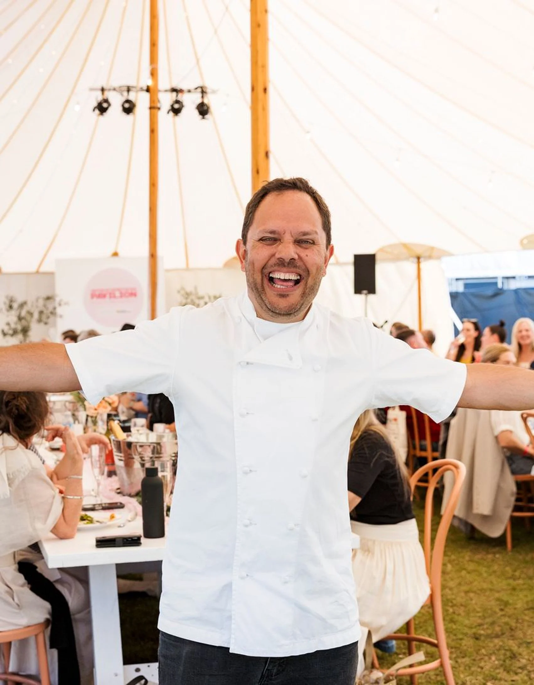 Happy man in a chef's coat with arms outstretched at a celebratory event under a large white tent.