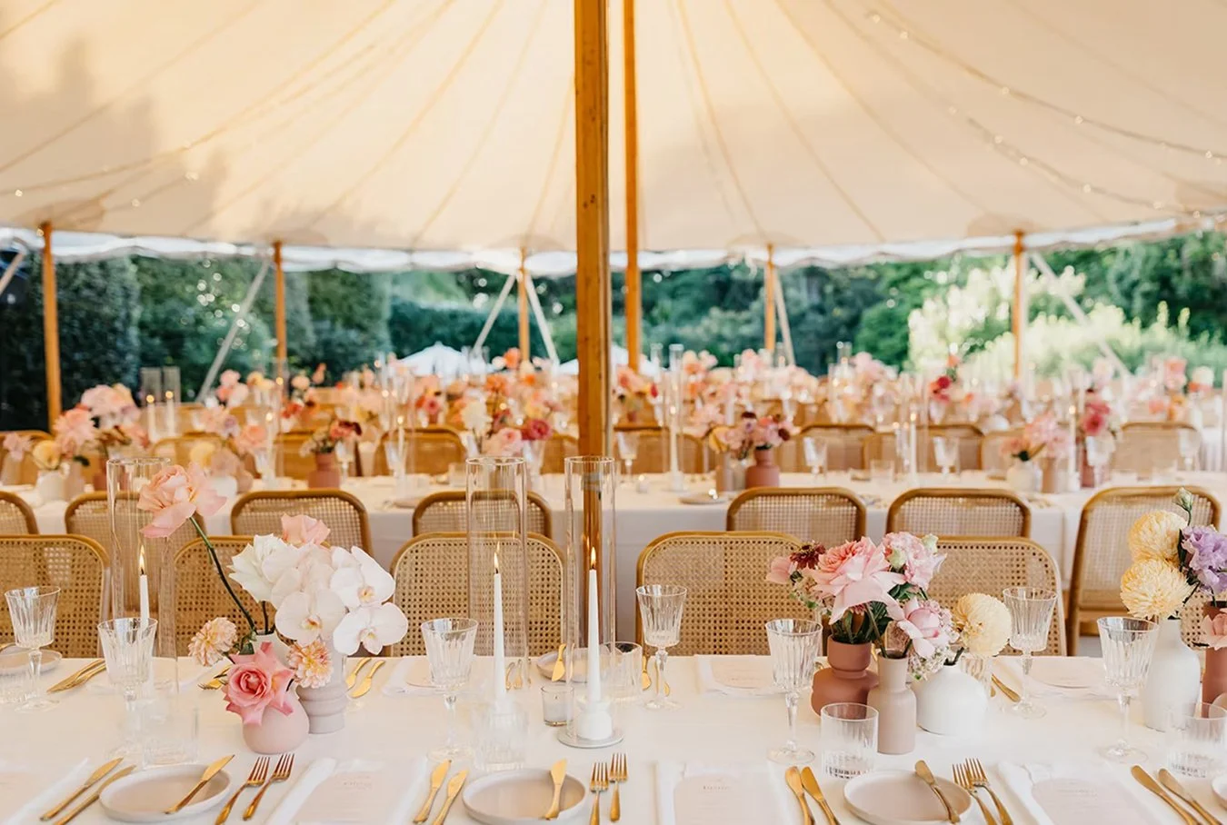 Elegant outdoor wedding reception under a large white tent with tables decorated with pink and white flowers in vases, gold flatware, and glassware.