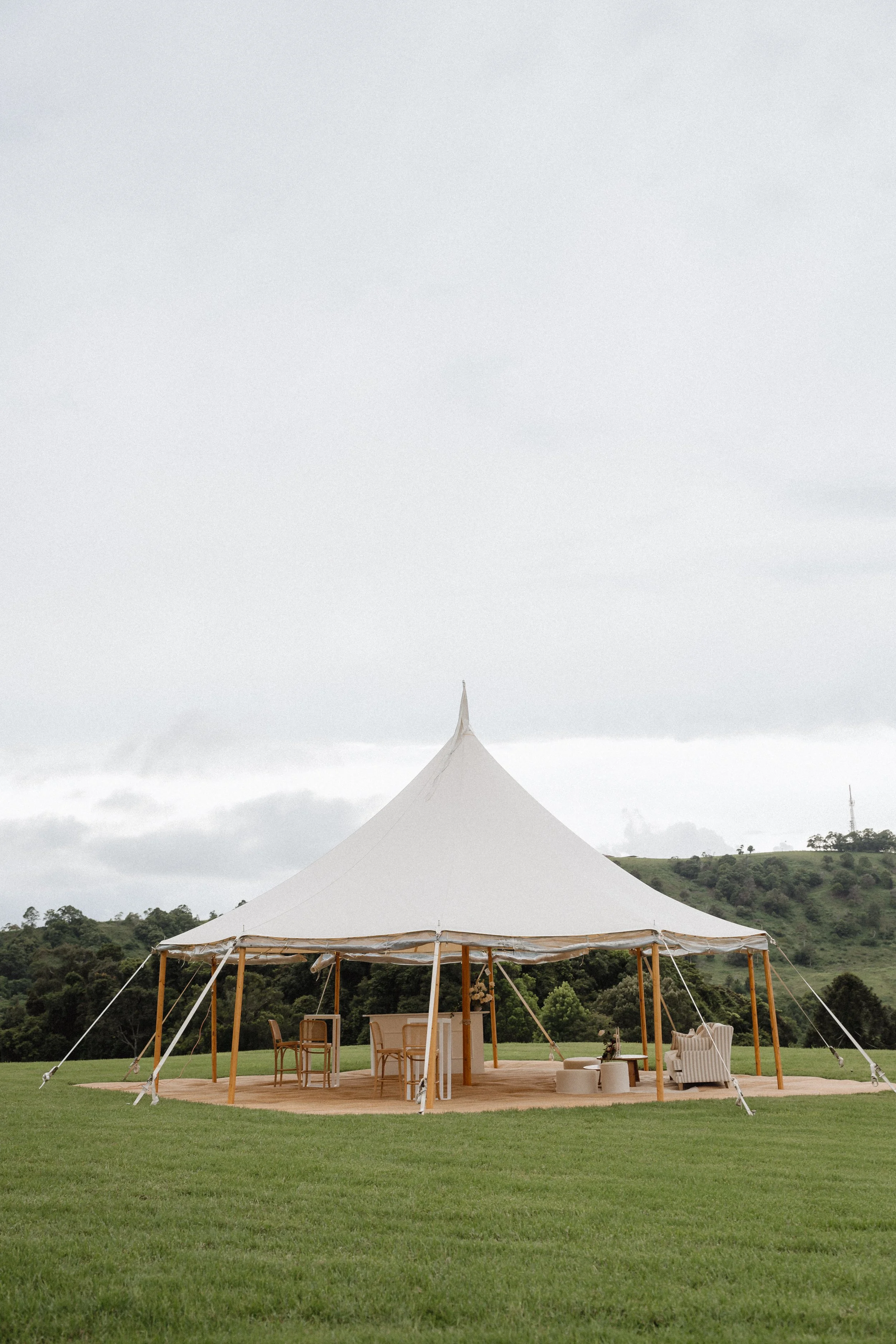 A large white tent set up on a grassy field with wooden poles and stakes, with outdoor furniture inside, and green hills in the background under a cloudy sky.