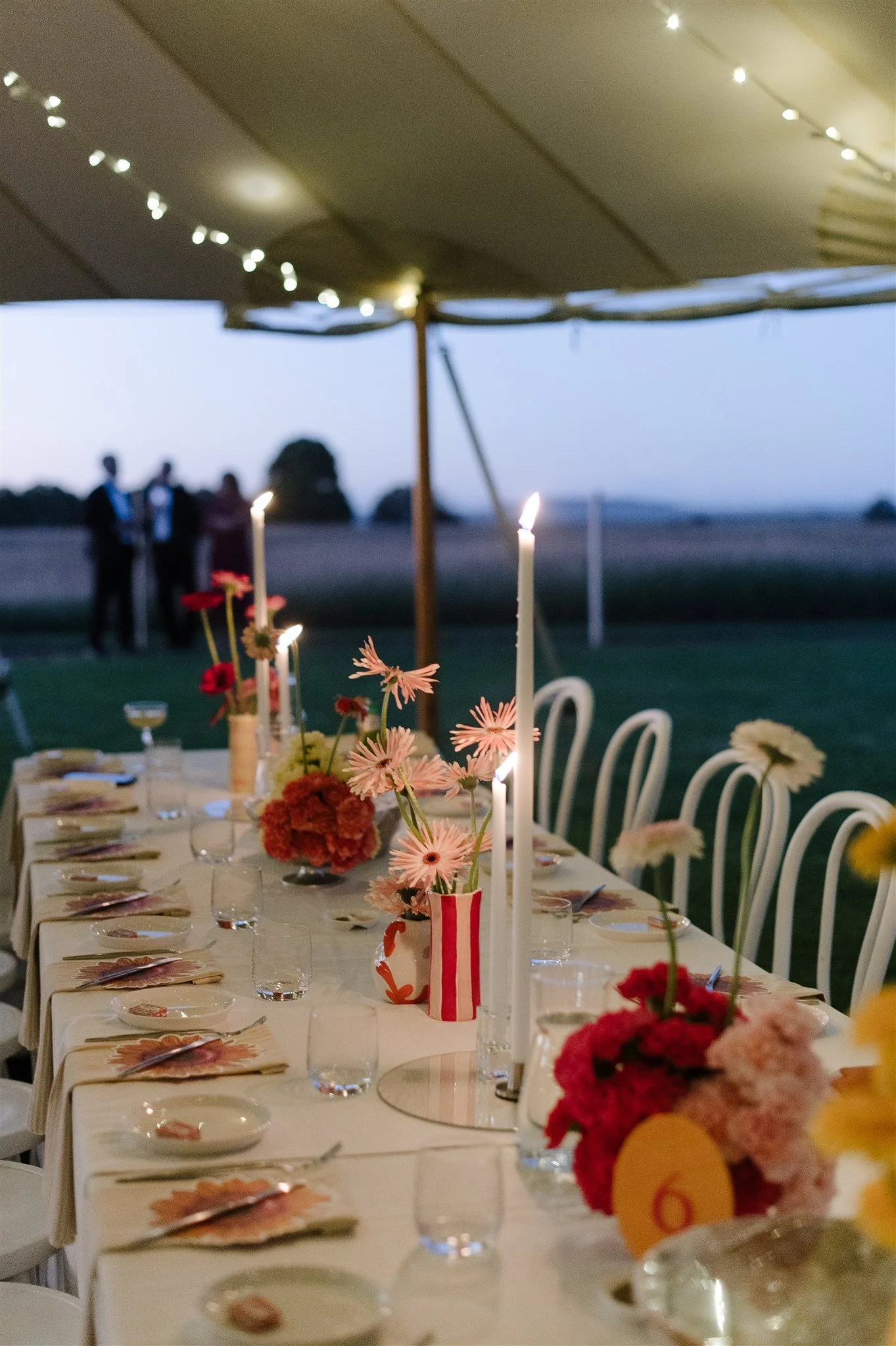 A long outdoor dining table set for a celebration, decorated with colorful flowers in vases, white candles, and place settings, under a canopy at dusk with a view of a field and trees in the background, and a group of people standing in the distance.