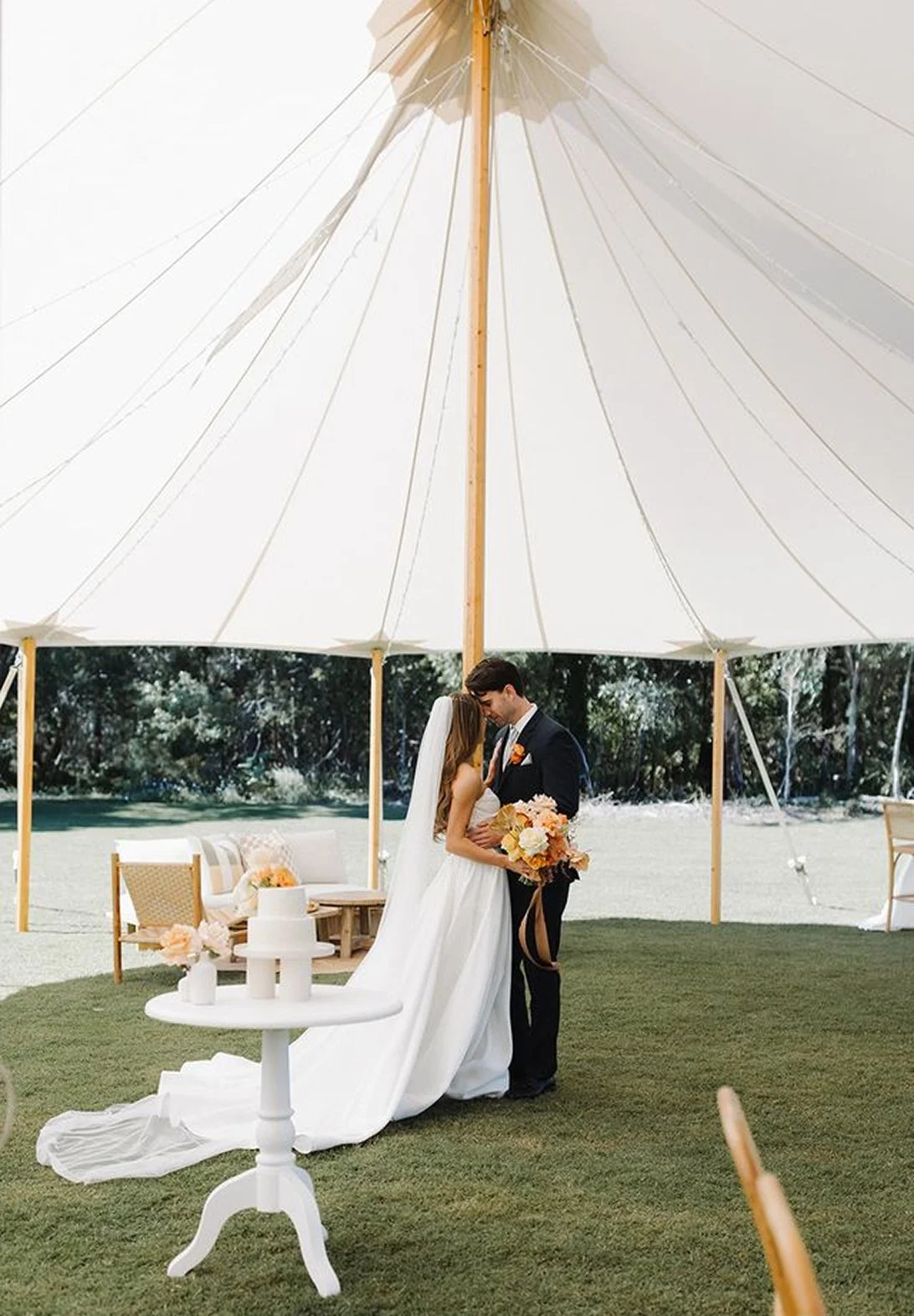 A bride and groom standing close together under a large white canopy tent on a grassy outdoor area, holding hands and leaning their foreheads together during their wedding ceremony. The bride is wearing a long white wedding dress and holding a bouquet of peach, cream, and pink flowers. The groom is dressed in a black suit with a white shirt and a peach-colored boutonniere. There are chairs and a small table decorated with flowers in the background.