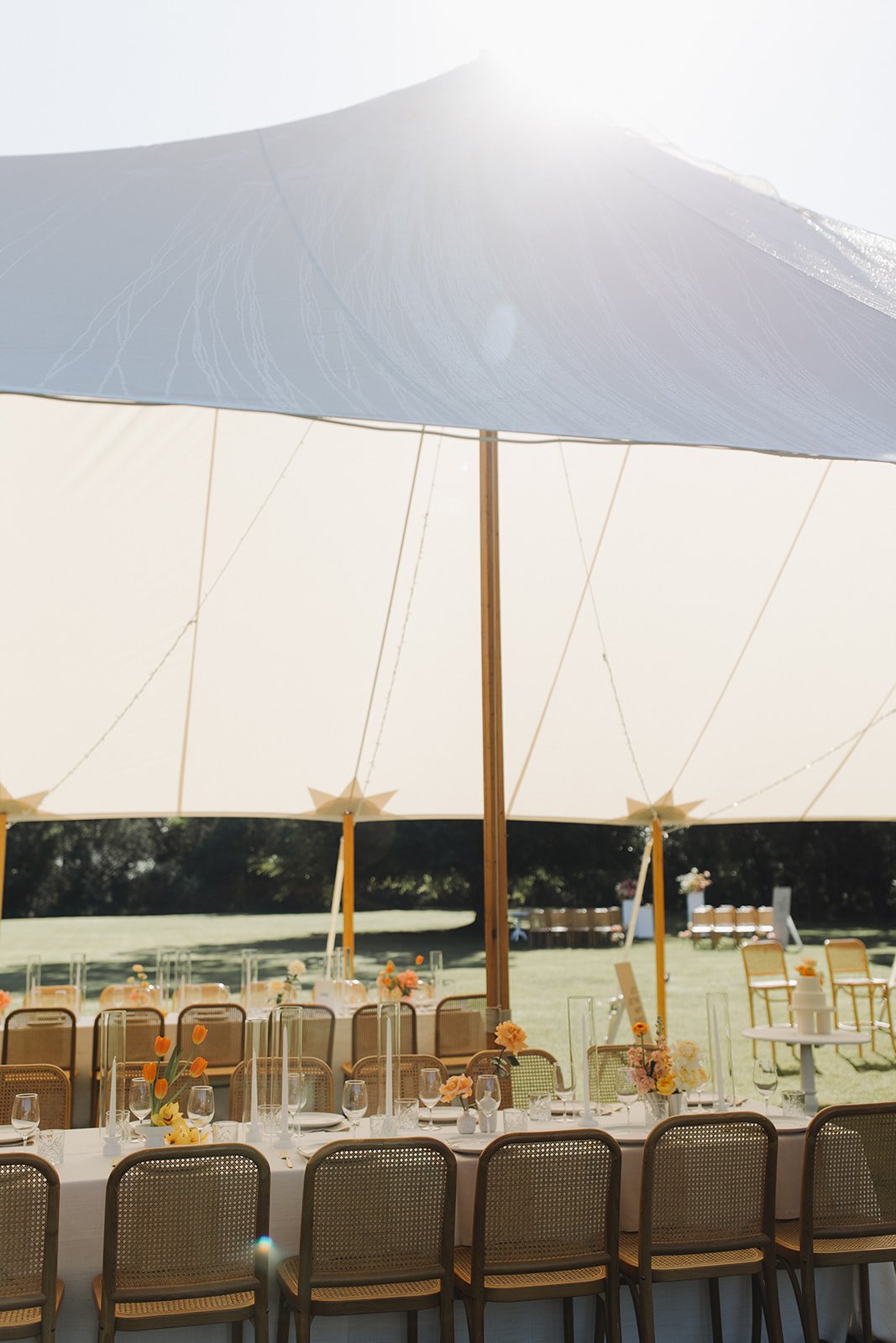Outdoor event setup with a long table, chairs, and floral decorations under a large white canopy tent on a sunny day.