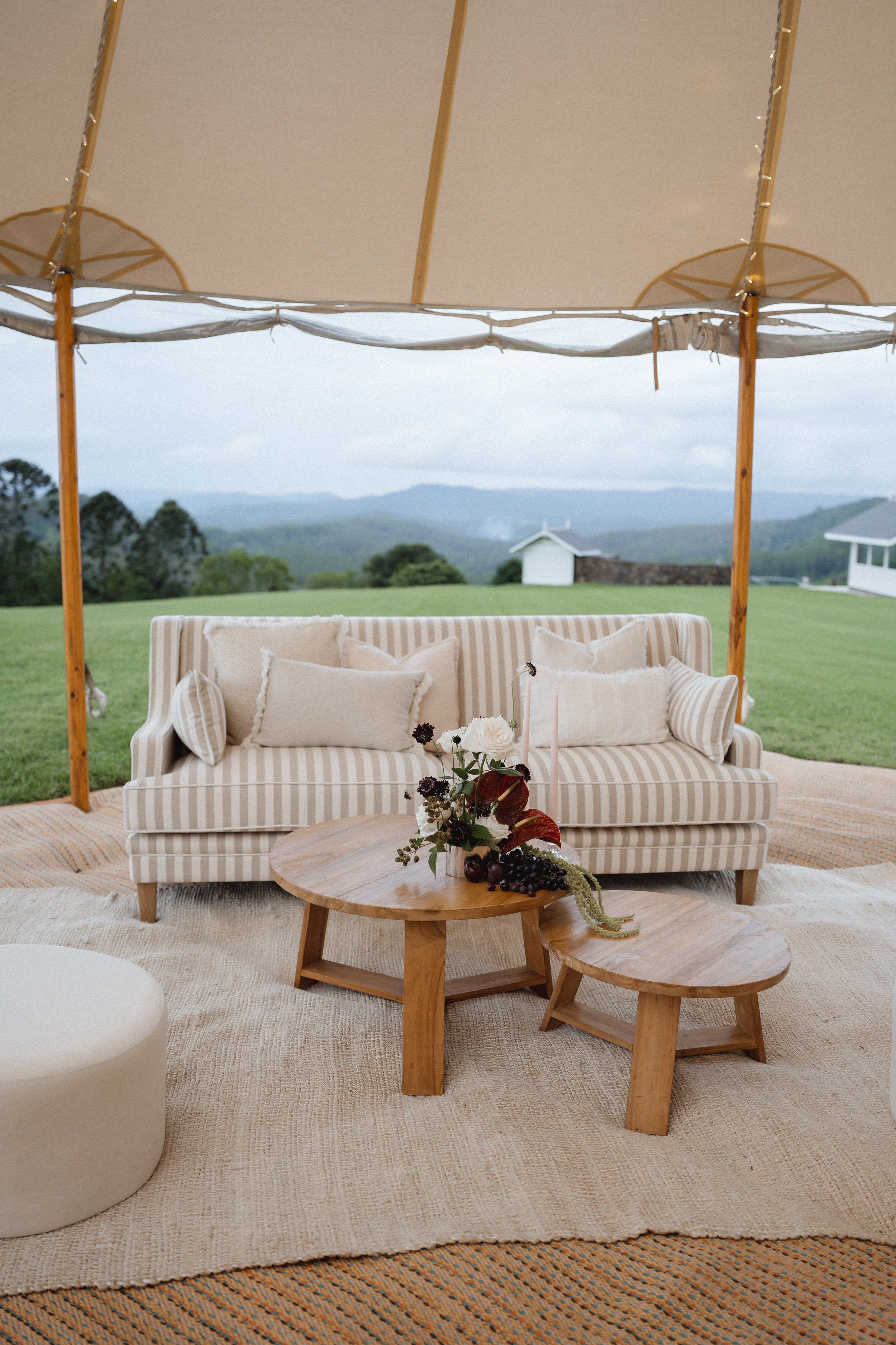 Outdoor event setup with striped sofa, wooden coffee tables and floral centerpiece, under a canopy with views of green hills and distant mountains.