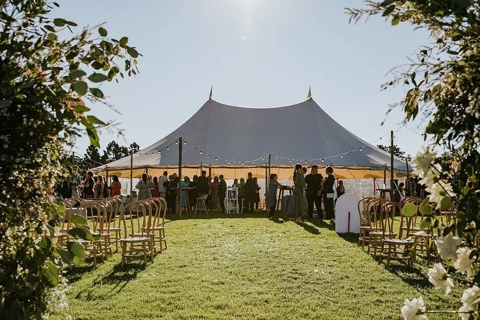 Outdoor event with a large white tent, people gathered inside and around, trees and chairs in the foreground, sunny day.