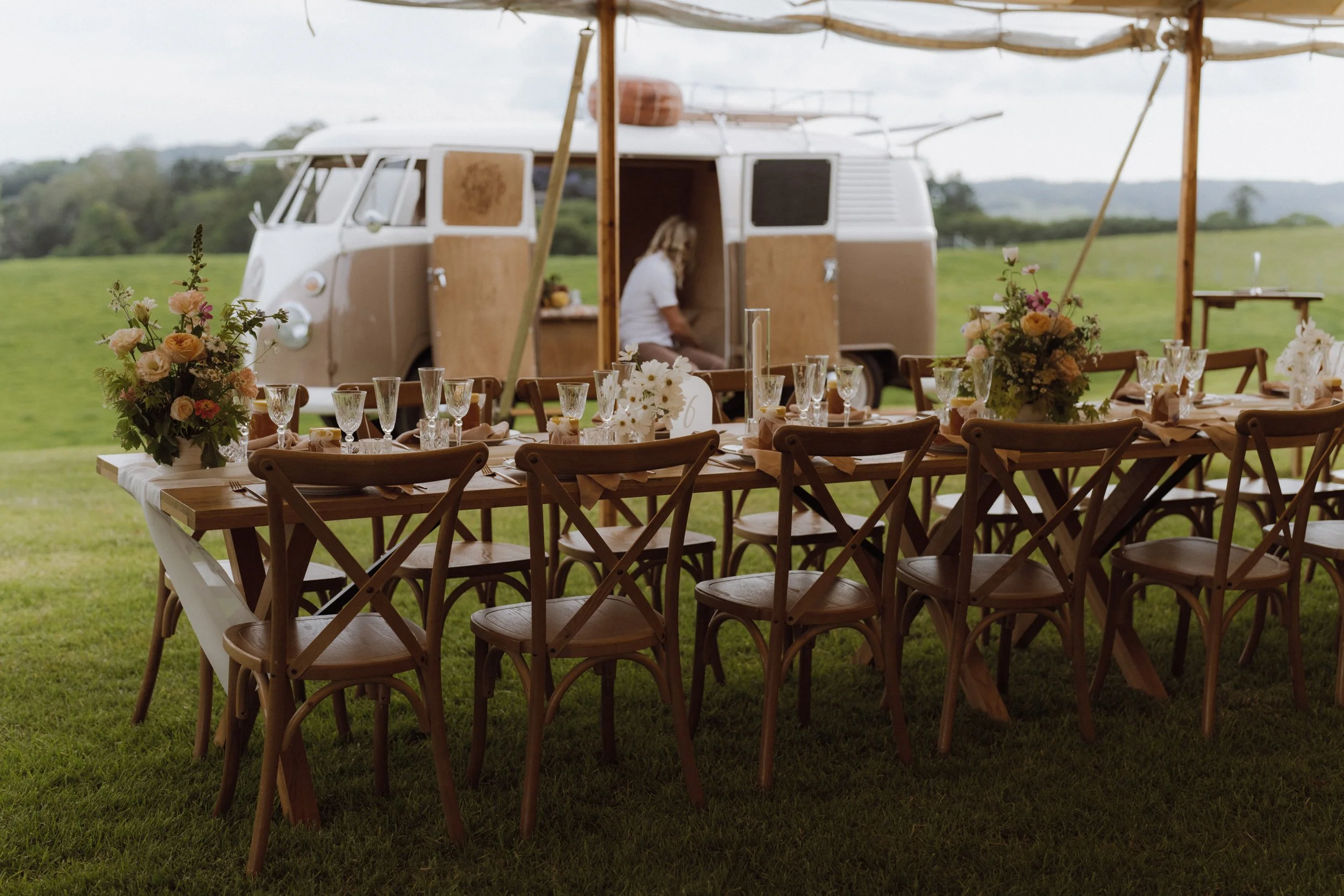 Outdoor dining setup with a long wooden table, chairs, and floral centerpieces, with a vintage camper van in the background and a person sitting inside the van.