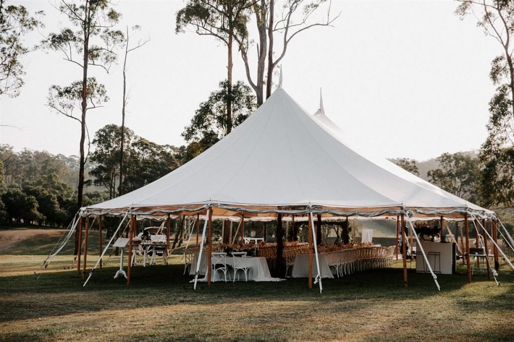 Large white event tent set up outdoors on grass with tall trees and cloudy sky in background, decorated for a gathering or celebration.