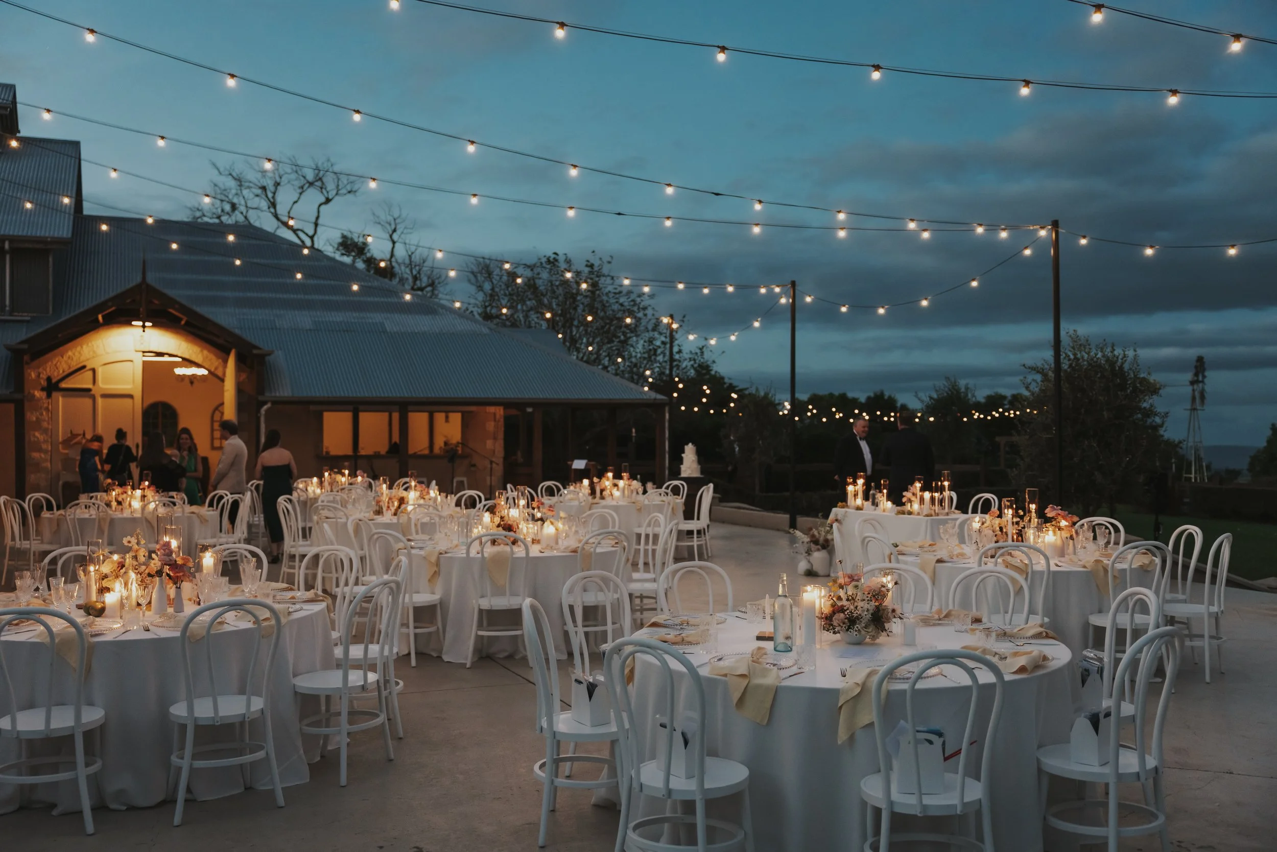 Outdoor wedding reception setup at dusk with round tables draped in white tablecloths, decorated with floral centerpieces, candles, and tableware. String lights hang overhead, and people in formal attire are mingling near a rustic building with a metal roof.