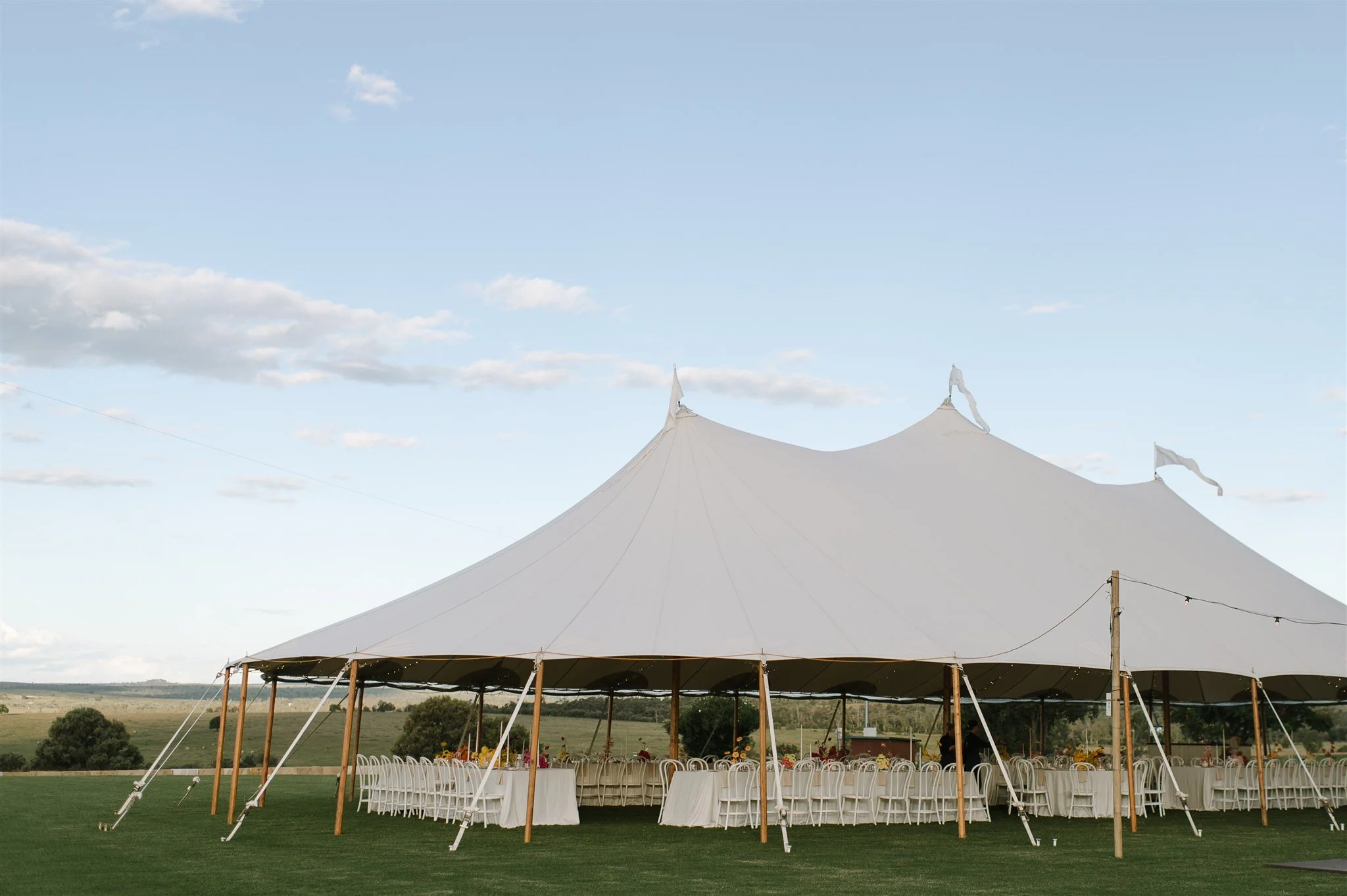 Large outdoor event tent with white fabric and wooden poles, set up on a grassy field with tables and chairs underneath, during daytime with partly cloudy sky.