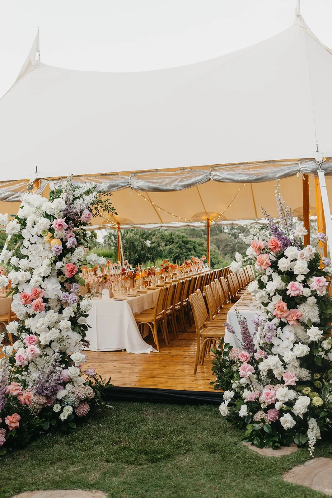 A wedding reception tent with long dining tables decorated with floral arrangements and string lights, viewed from outside on grass and wood flooring.