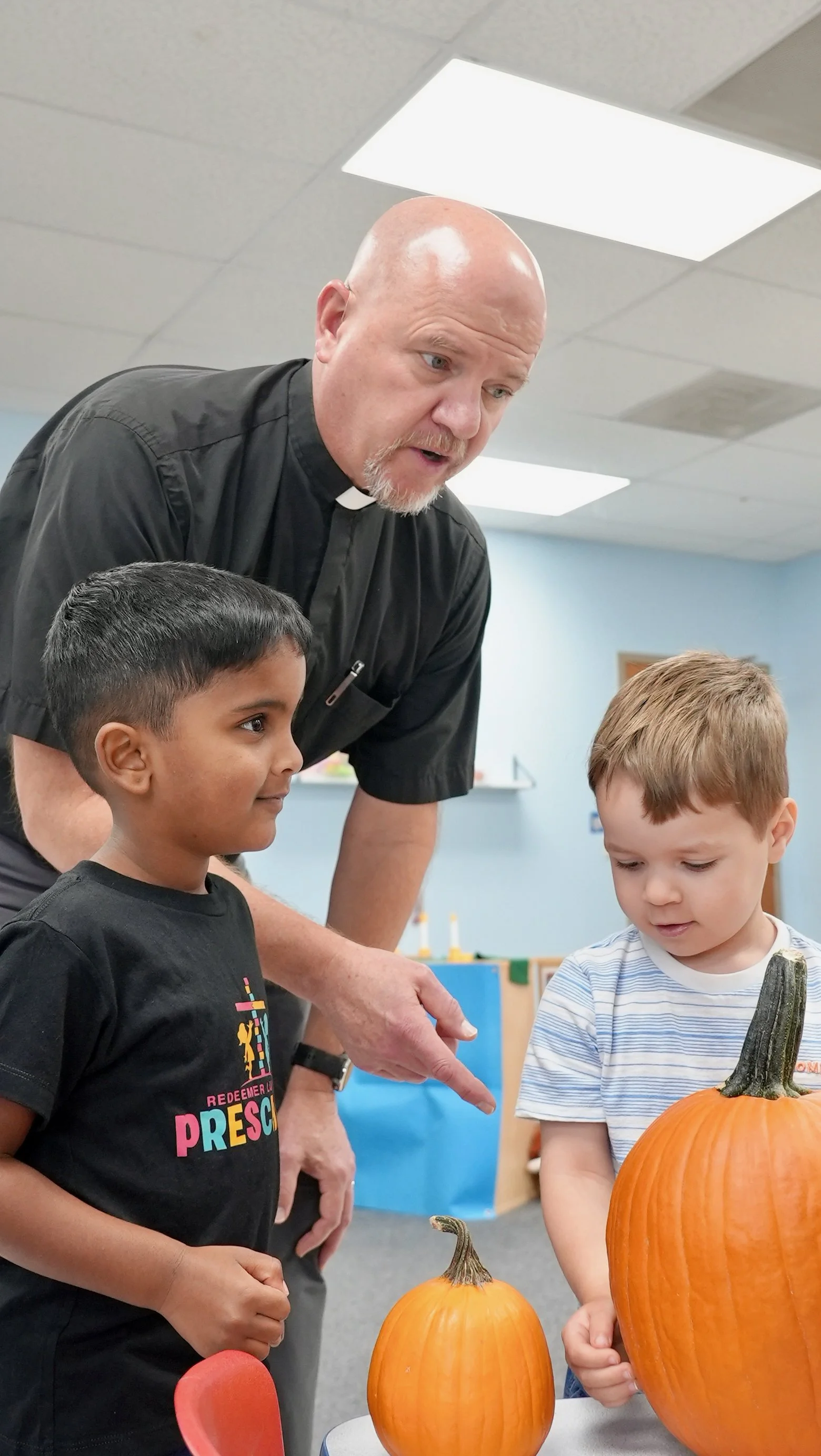 Pastor Kahle at Redeemer Lutheran Preschool in Olathe, Kansas, showing two young boys pumpkins in a classroom setting during a learning activity.