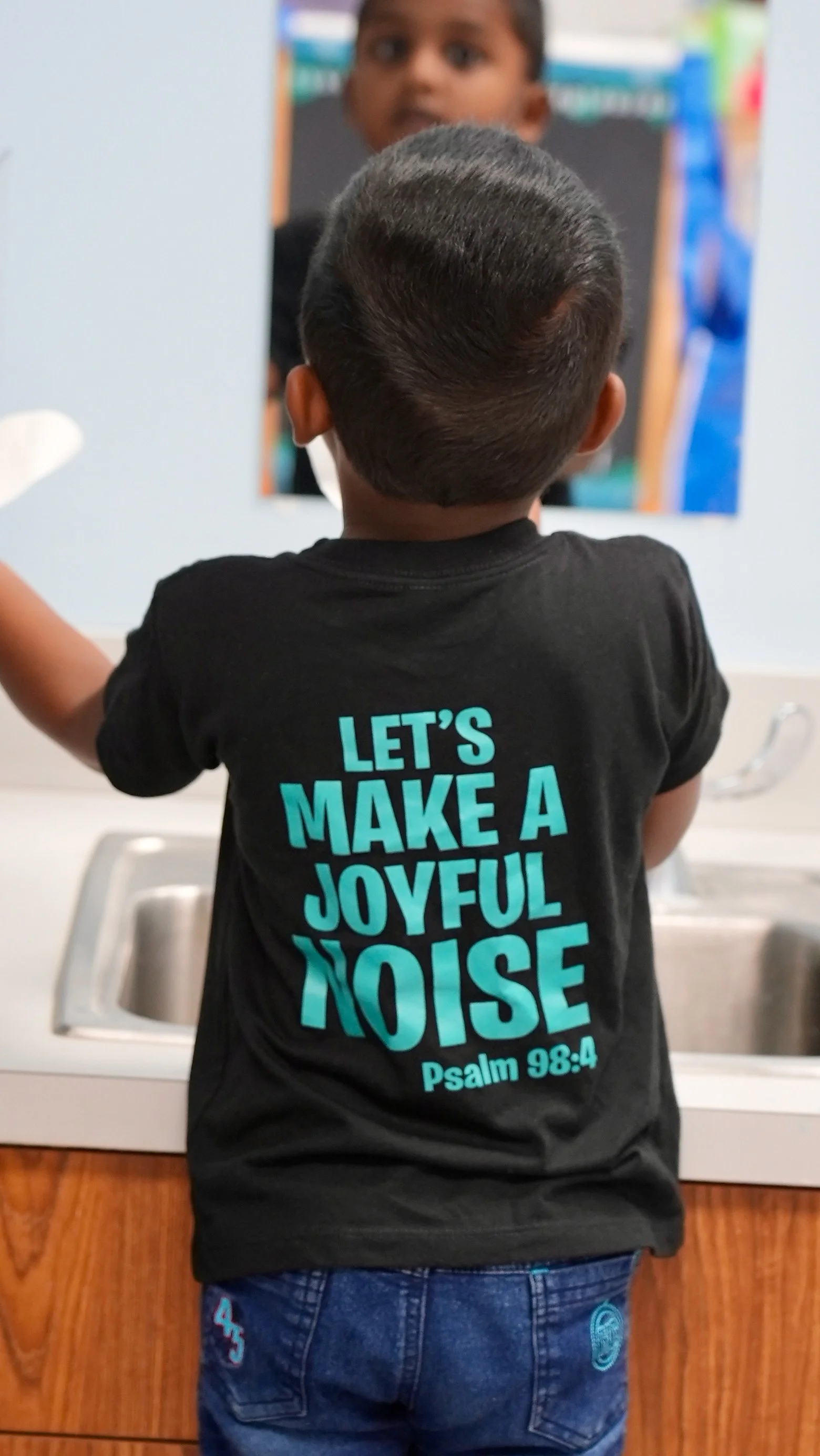 A young boy at Redeemer Lutheran Preschool standing in front of a mirror, wearing a black T-shirt with the text ‘Let’s Make a Joyful Noise, Psalm 98:4,’ reflecting his face and joyfully engaging in a classroom activity.